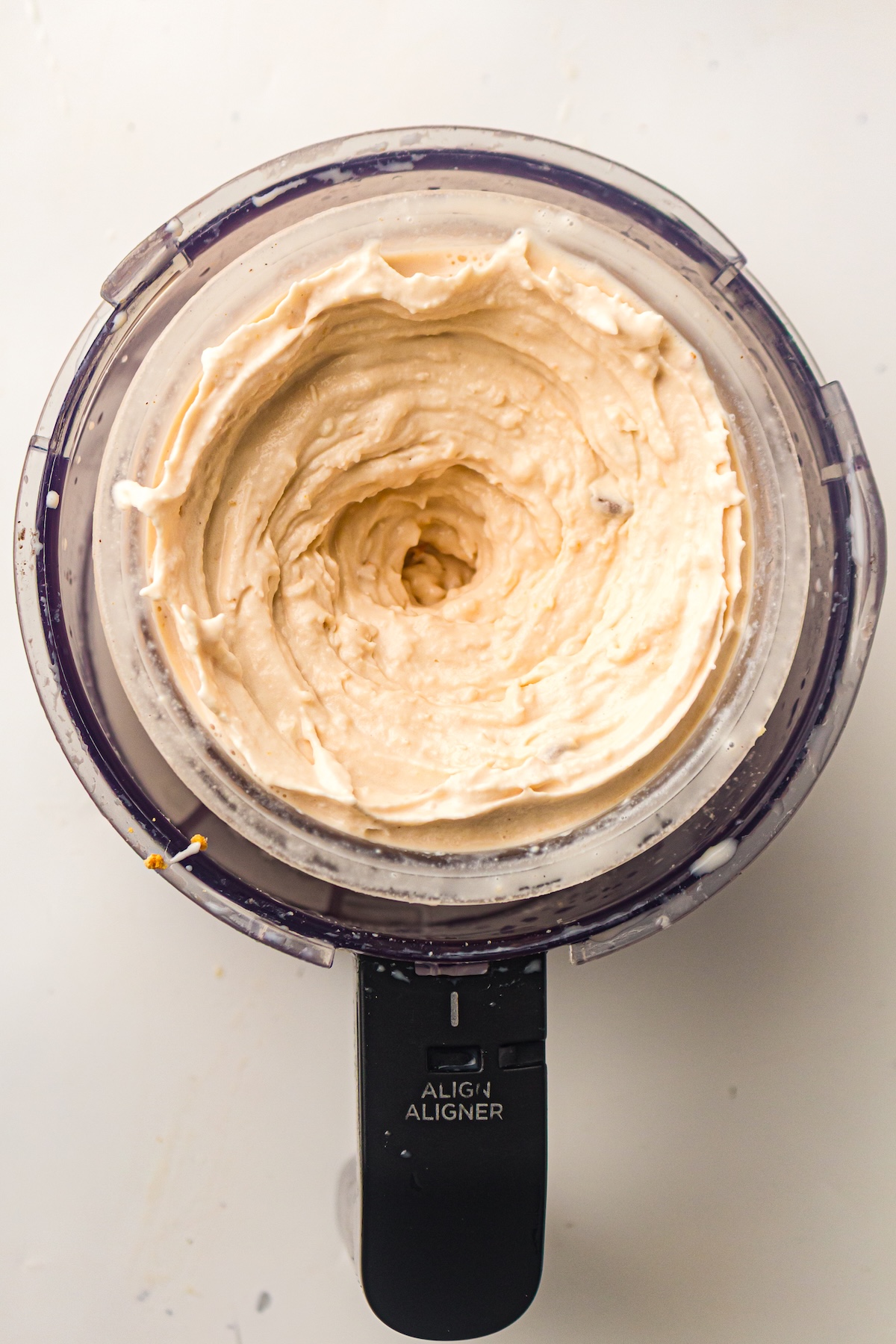 Overhead view of a clear container from a kitchen appliance, filled with a thick, light beige creamy mixture with swirls and small dark flecks, featuring a black handle labeled 'ALIGN ALIGNER,' set on a white background.