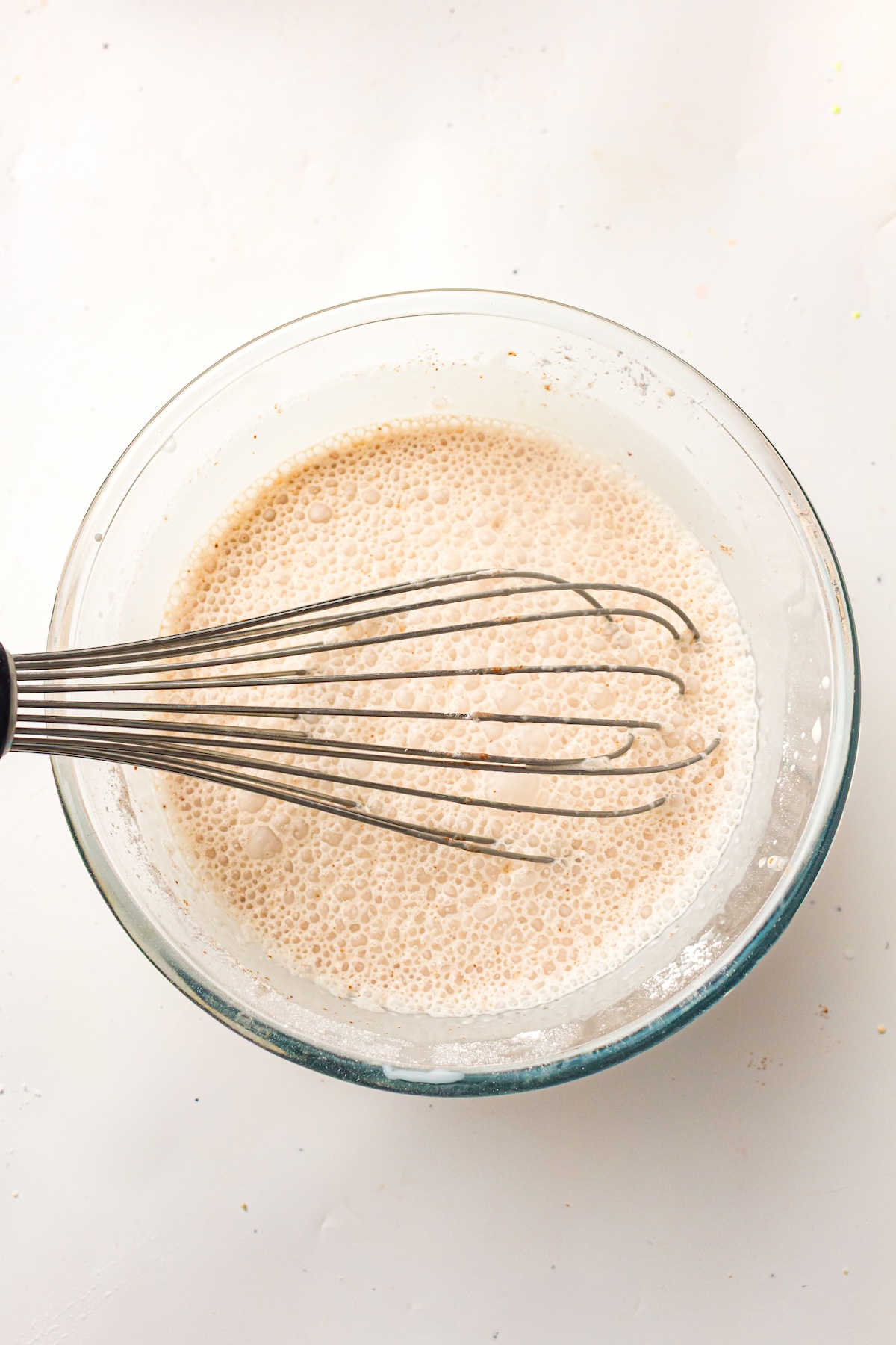 Top-down view of a clear glass bowl with a light brown frothy liquid and a metal whisk partially submerged, set on a light-colored surface.