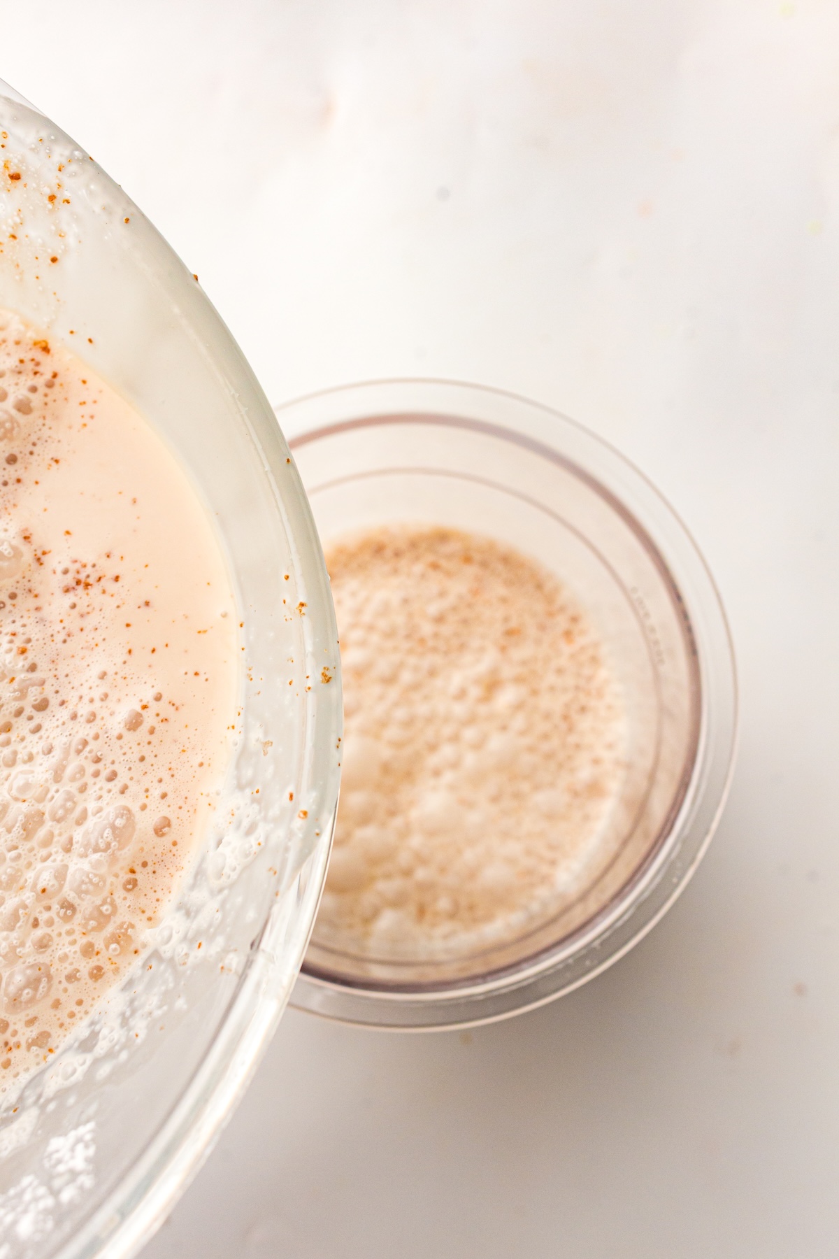 Close-up of a light beige foamy liquid being poured from one clear glass bowl into another, with small reddish-brown specks visible in the foam, set against a plain white background.