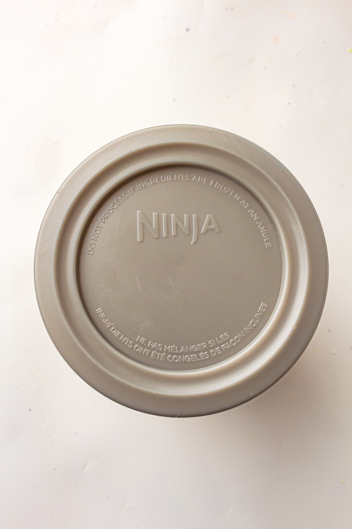 Overhead view of a gray circular lid from a Ninja appliance, featuring embossed text with safety instructions in English and French, set on a light speckled surface.