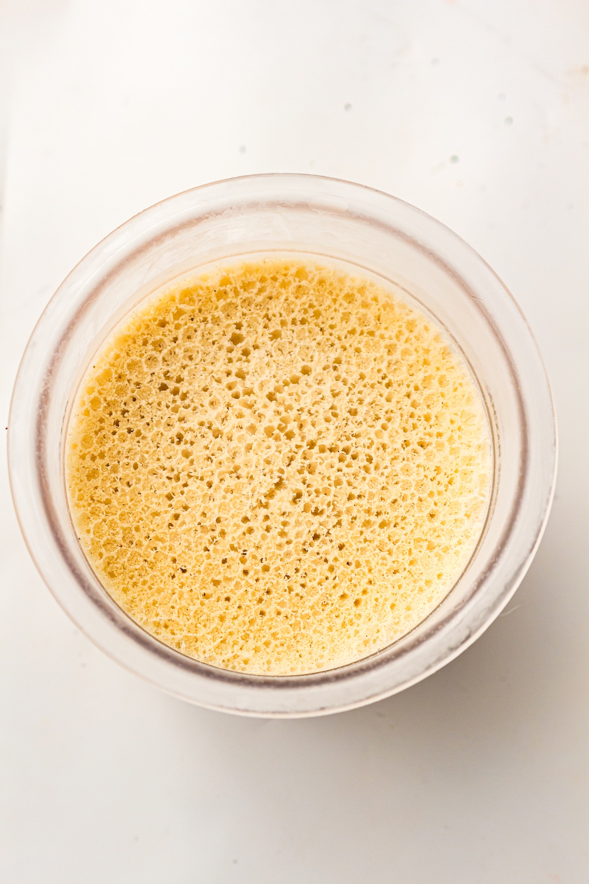 Top-down view of a clear container filled with a light yellow frothy or porous substance, with small dark specks scattered across the surface, set on a plain white background.