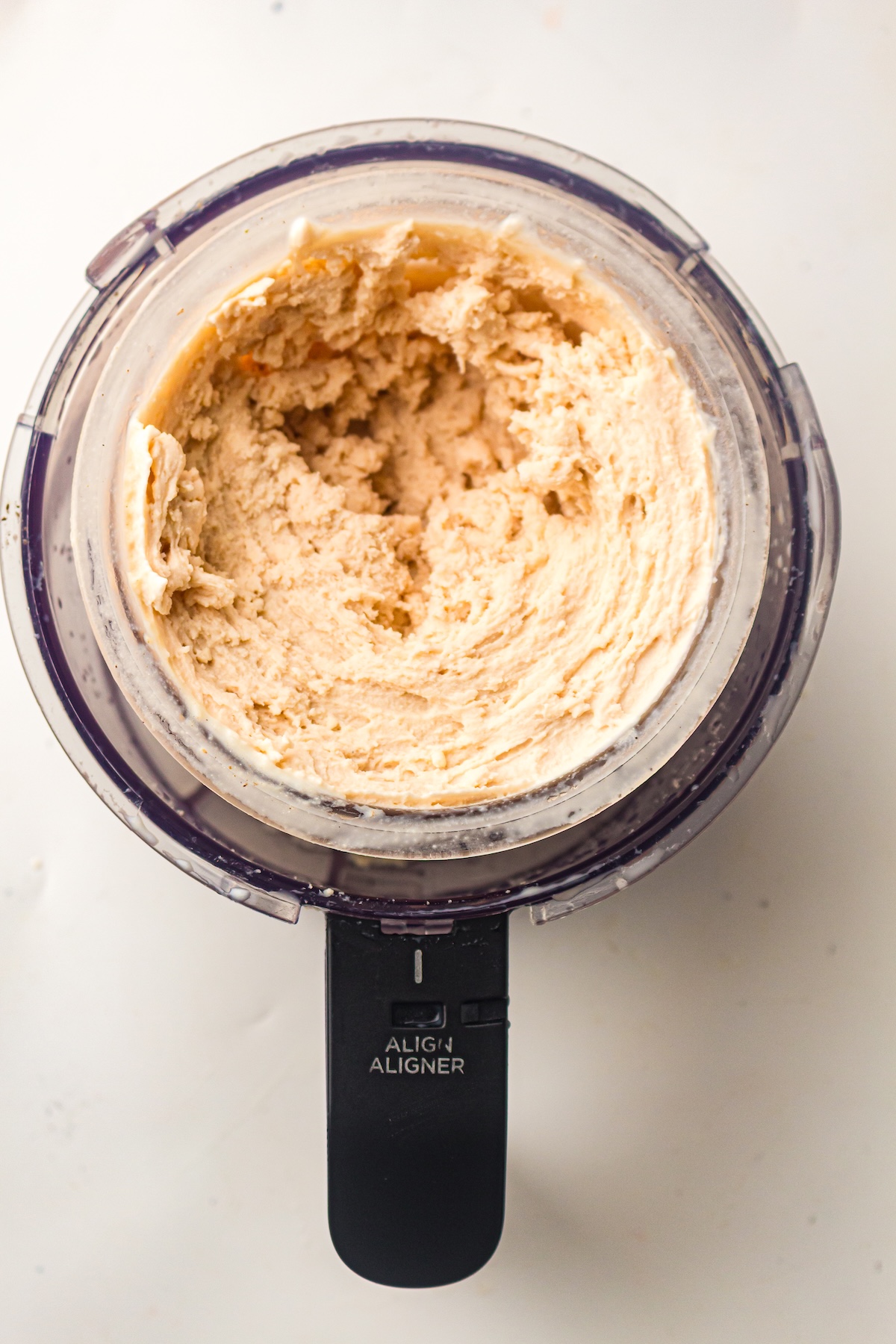 Overhead view of a transparent container from a Ninja appliance, filled with a light brown creamy mixture with swirls and peaks, featuring a black handle labeled 'ALIGN ALIGNER,' set on a white countertop.