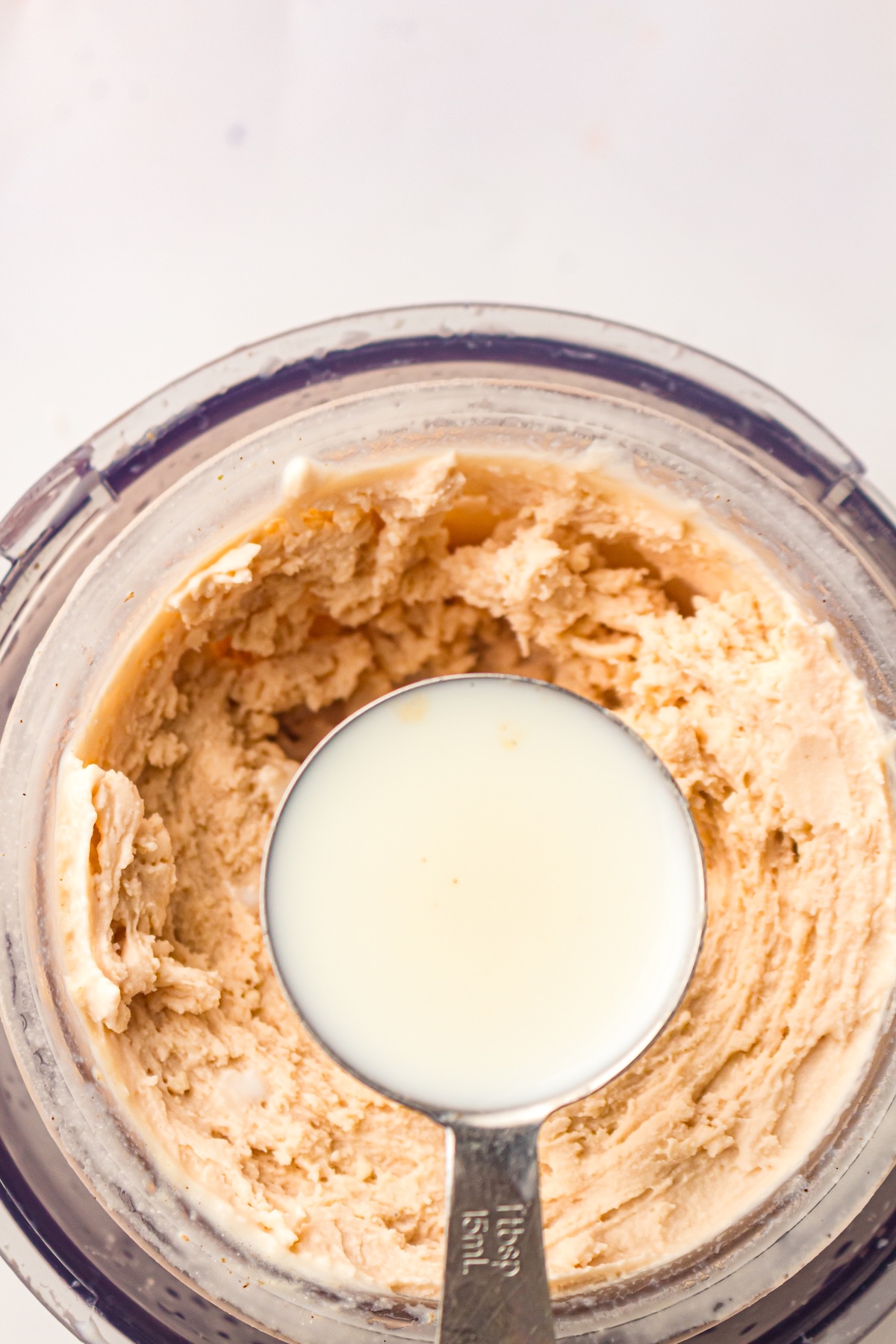 Top-down view of a clear container filled with a thick, light brown textured mixture, with a silver measuring spoon marked '1tbsp 15mL' resting in the mixture and holding a small amount of off-white liquid, set on a light background.