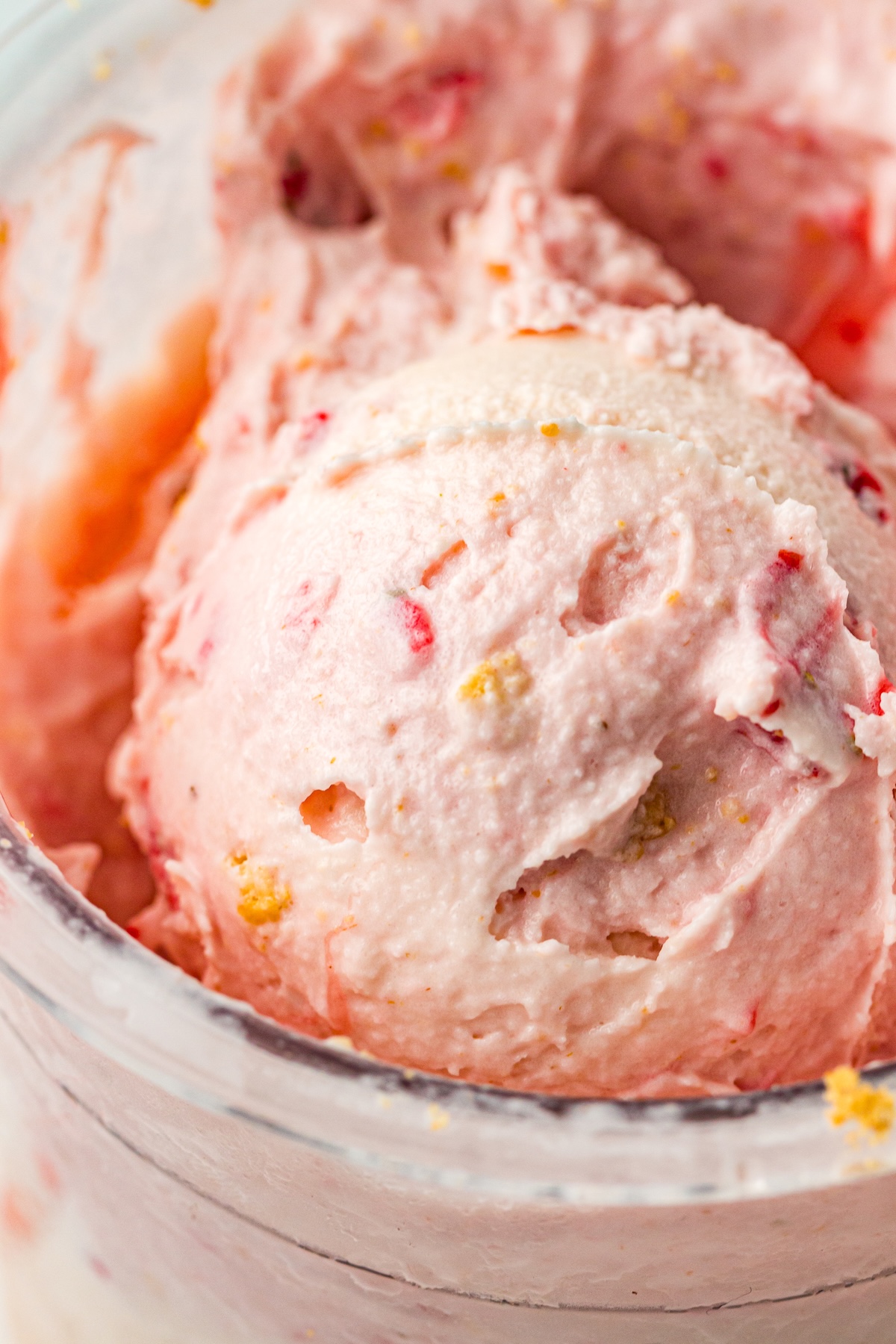 Close-up of a scoop of light pink strawberry cheesecake ice cream in a clear container, featuring visible red fruit pieces and yellowish flecks, set against a softly blurred background.