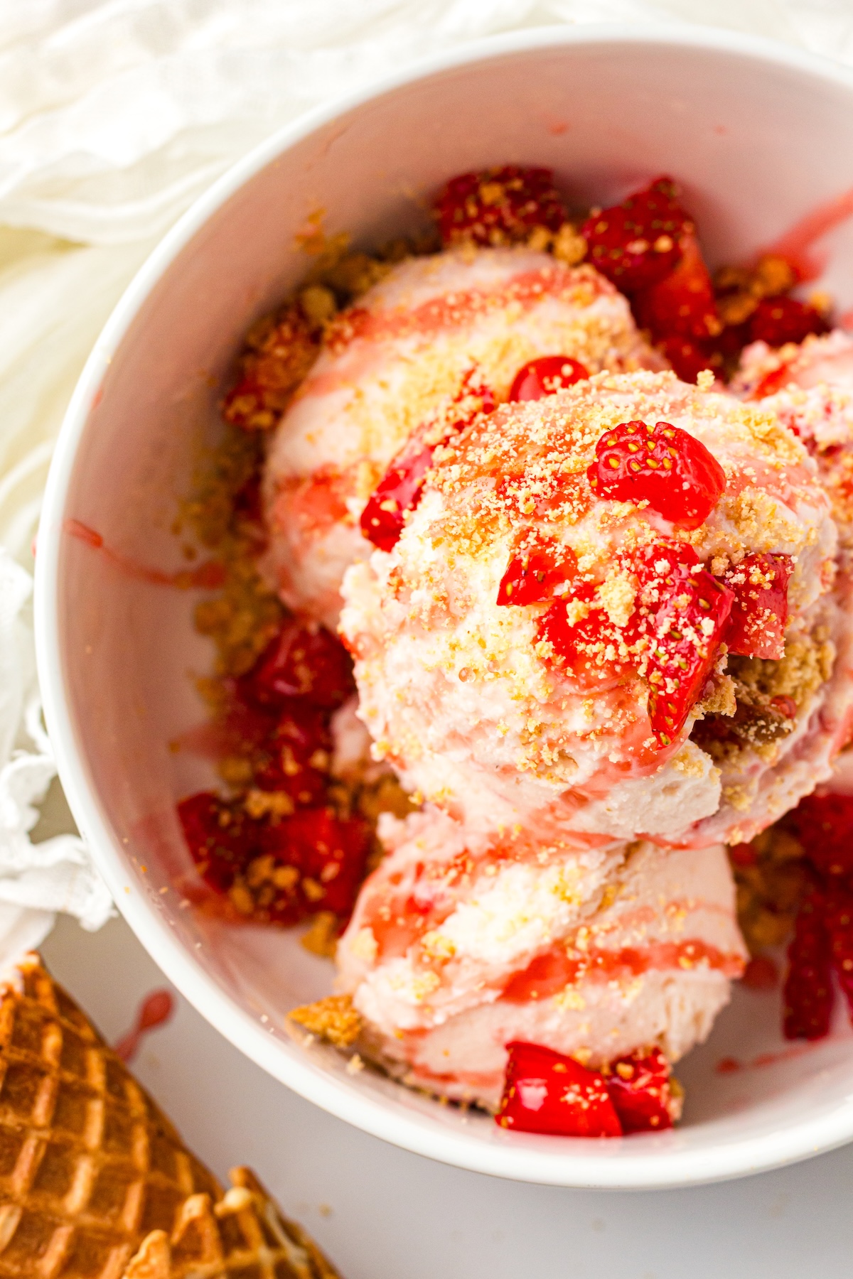 Close-up of a white bowl filled with scoops of light pink strawberry or cherry ice cream, garnished with bright red fruit pieces, golden crumbly topping, and drizzles of red syrup. A waffle cone is partially visible in the foreground.
