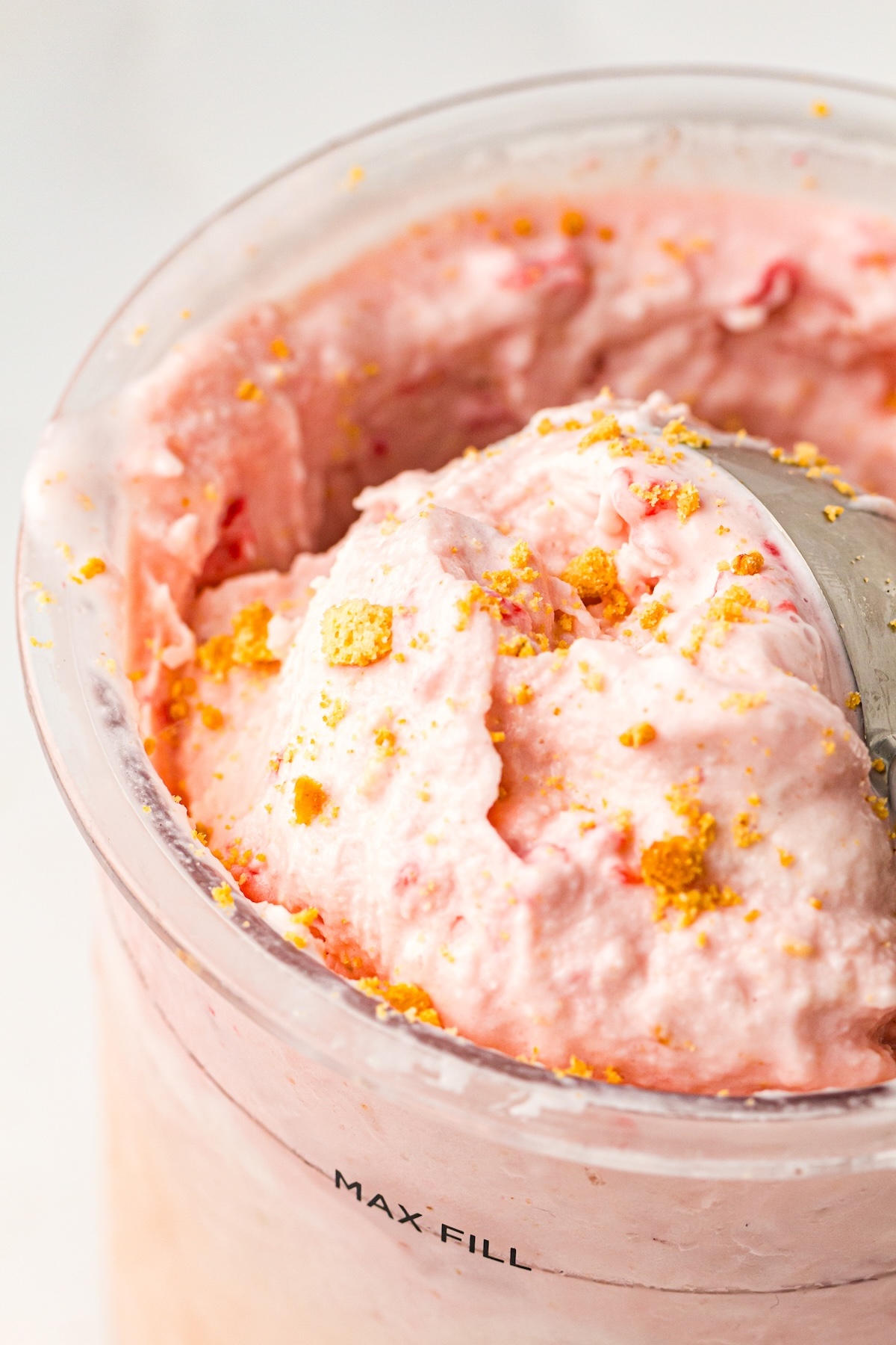 Close-up of light pink strawberry cheesecake ice cream in a clear container, topped with golden-brown crumbs, with a silver scoop partially visible, and 'MAX FILL' text printed on the side, set against a bright white background.