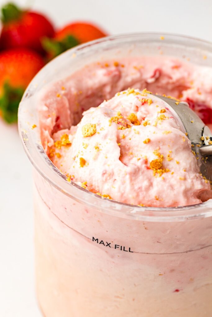 Top-down view of light pink creamy strawberry cheesecake ice cream in a clear container, topped with golden-brown crumbs, with a metal scooper partially submerged, and fresh strawberries blurred in the background.
