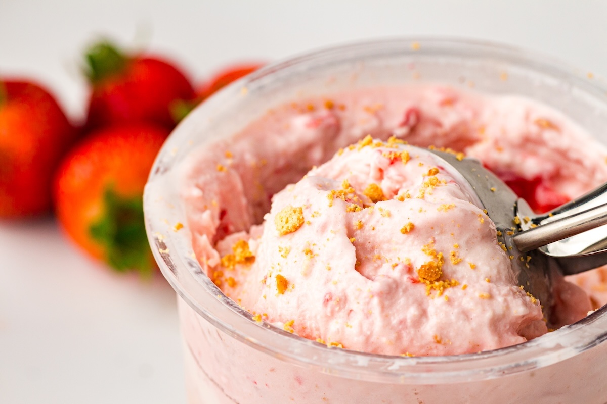 Close-up of light pink strawberry cheesecake ice cream in a clear container, garnished with golden crumbs, with a metal scoop partially submerged, and fresh strawberries blurred in the background.