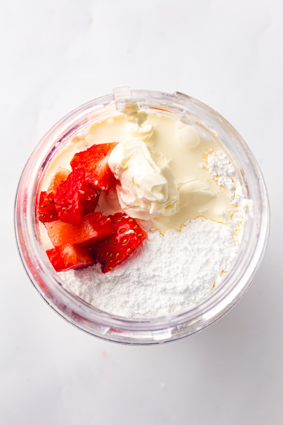 Top-down view of a blender cup containing fresh strawberry chunks, cream cheese, whipped cream, and powdered sugar, set against a clean white background.