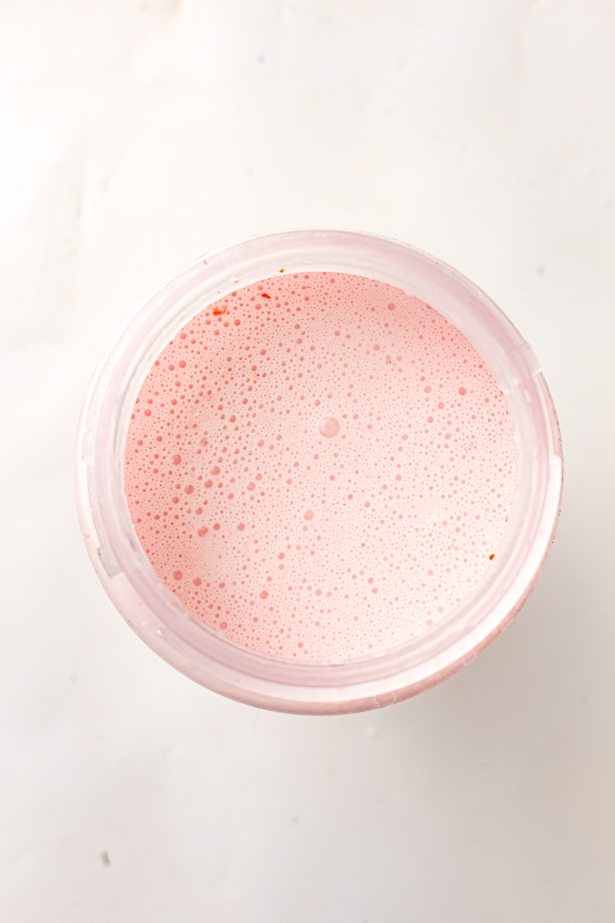 Close-up of a light pink foamy liquid with small reddish specks, contained in a translucent cup, set against a bright white background.