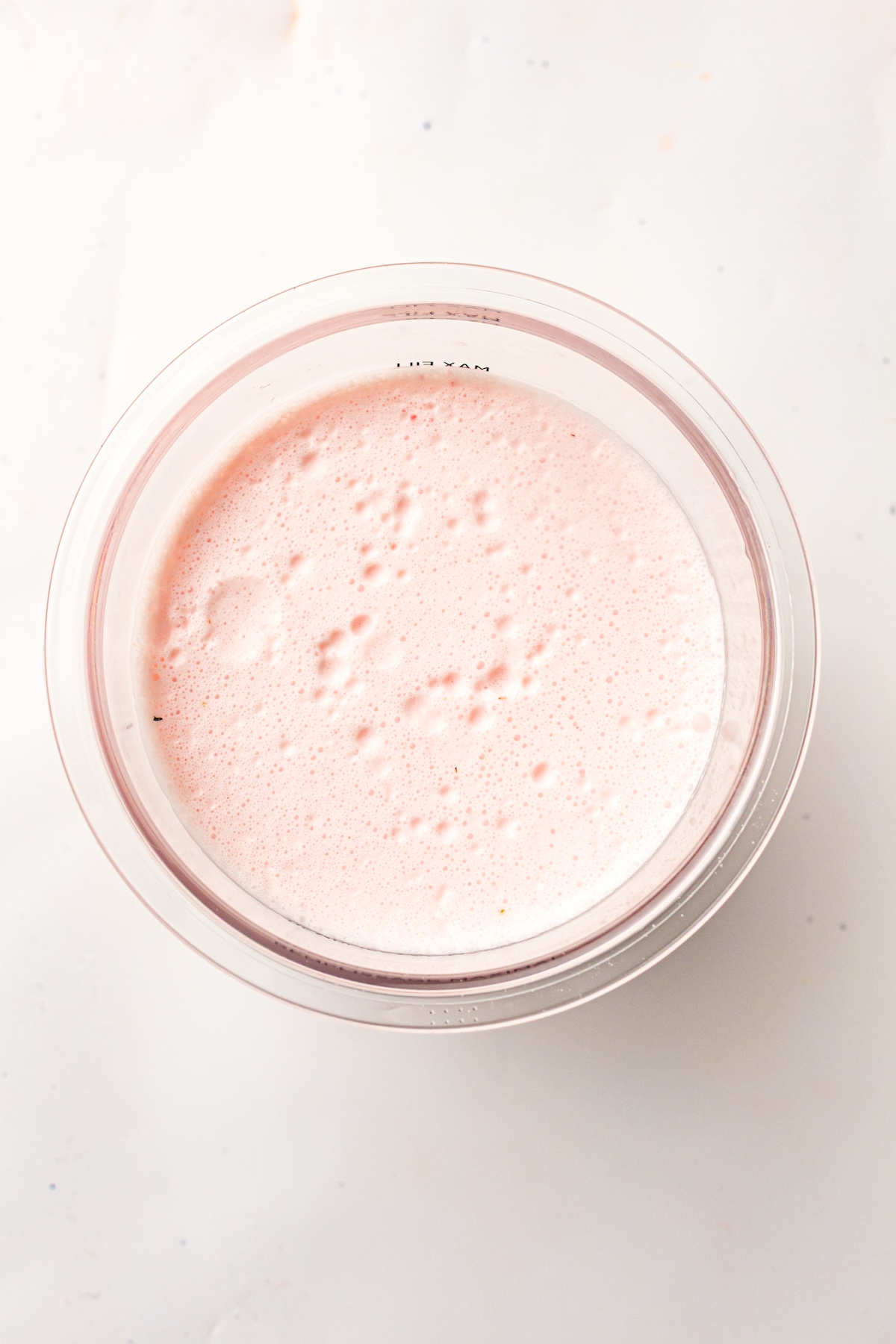 Overhead view of a clear glass bowl filled with light pink foamy liquid, with small bubbles and reddish specks on the surface, set against a light-colored textured background.