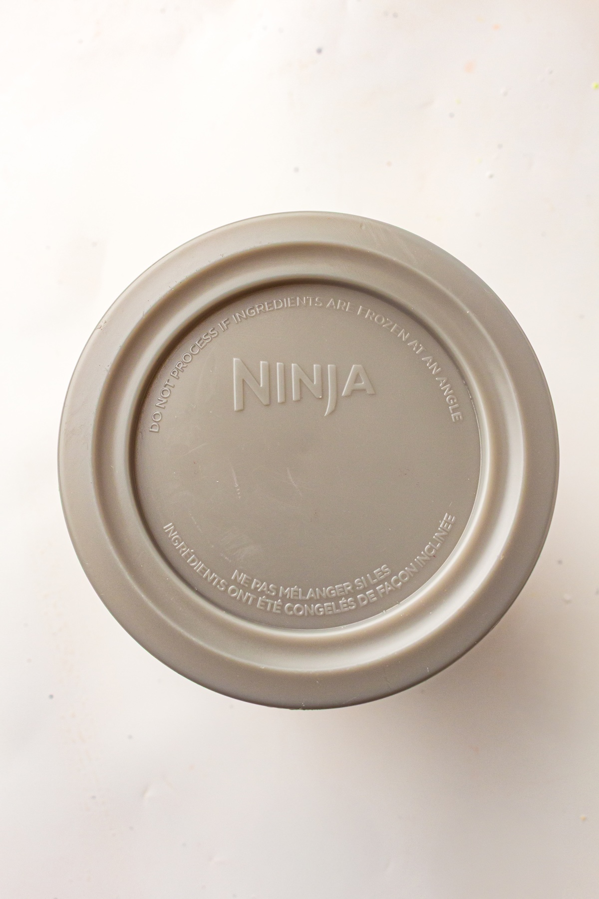 Close-up of a gray circular Ninja appliance lid with embossed text, including 'DO NOT PROCESS IF INGREDIENTS ARE FROZEN AT AN ANGLE' and 'NINJA,' set against a light background.