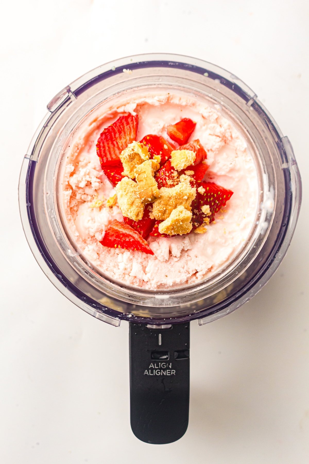 Overhead view of a clear container filled with light pink ice cream, topped with fresh strawberry pieces and golden biscuit crumbs, attached to a black base labeled 'ALIGN ALIGNER,' set against a light background.