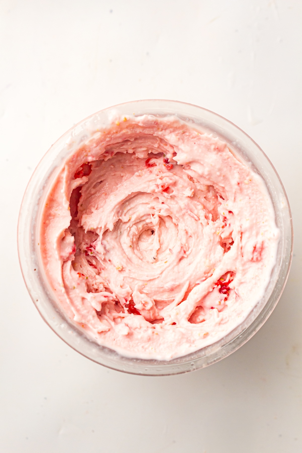 Close-up of a clear bowl containing light pink swirled ice cream with red fruit pieces, set against a plain light background, emphasizing the dessert's creamy texture and vibrant colors.