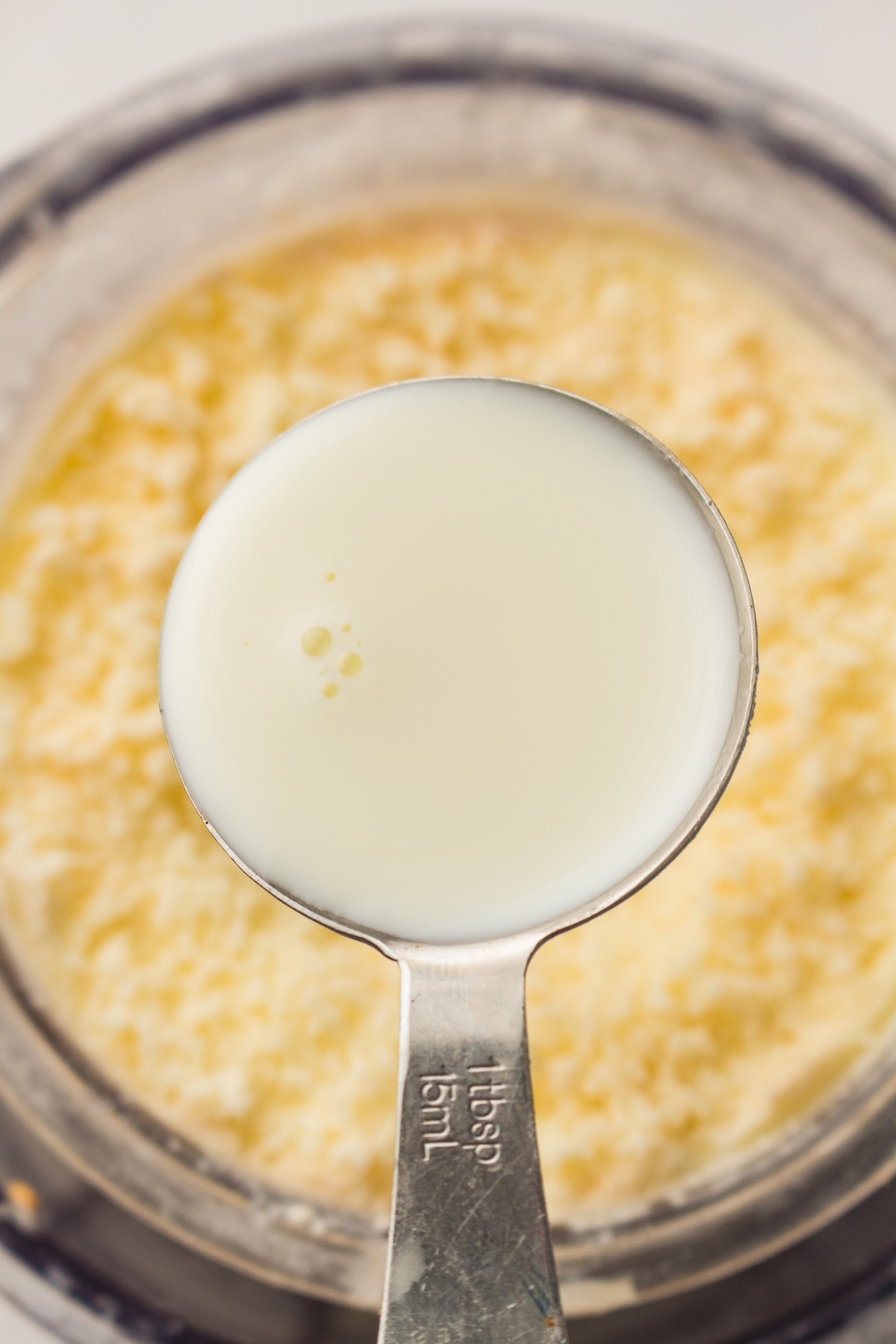 Close-up of a metallic measuring spoon filled with 1 tablespoon (15mL) of milky white liquid, held above a blurred container of yellow, granular substance, set against a light background.