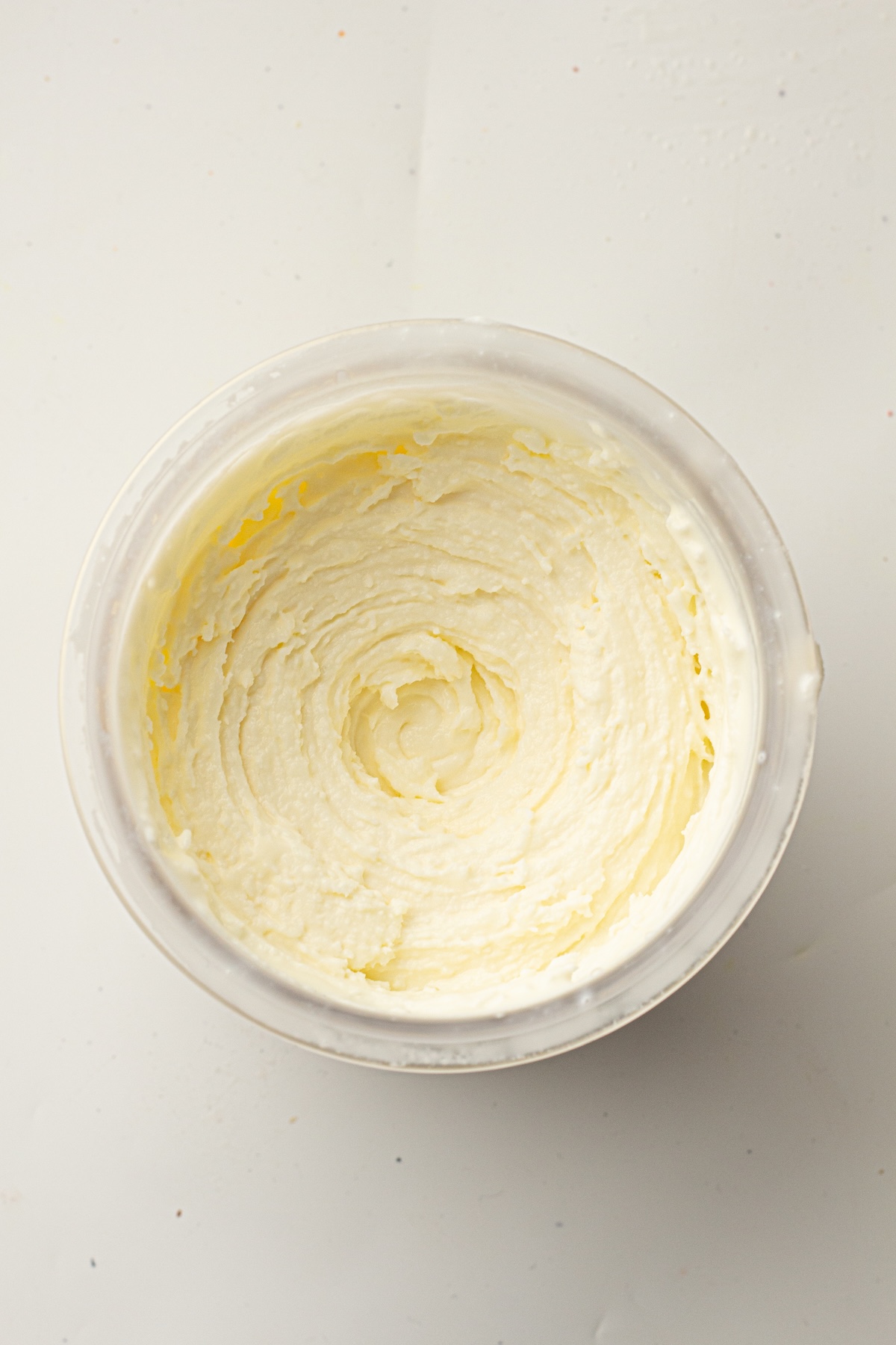 Overhead view of a clear plastic container filled with a thick, pale yellow creamy substance with swirled patterns, set on a plain light-colored background.
