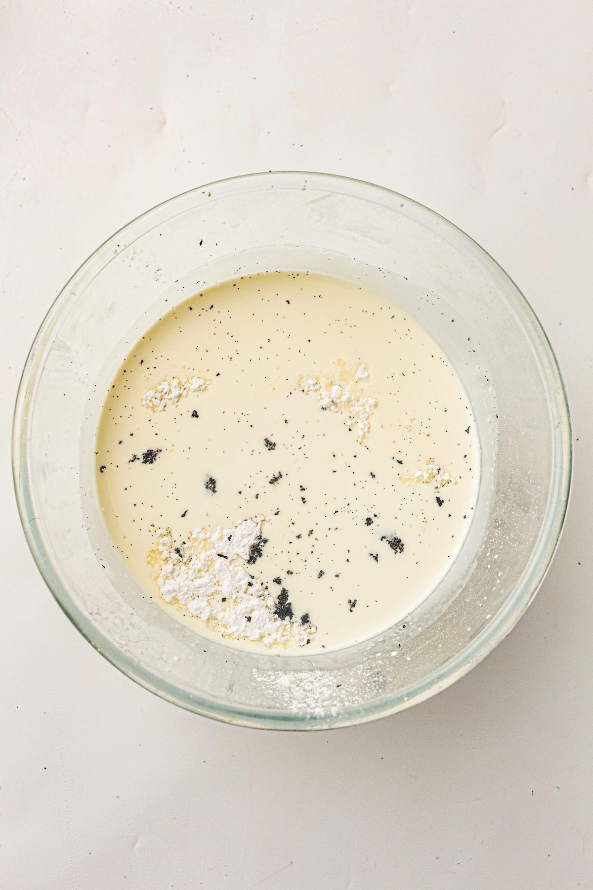 Overhead view of a clear glass bowl filled with a light yellow liquid, scattered with fine black specks, larger dark clumps, and piles of white powder, set on a light off-white background.