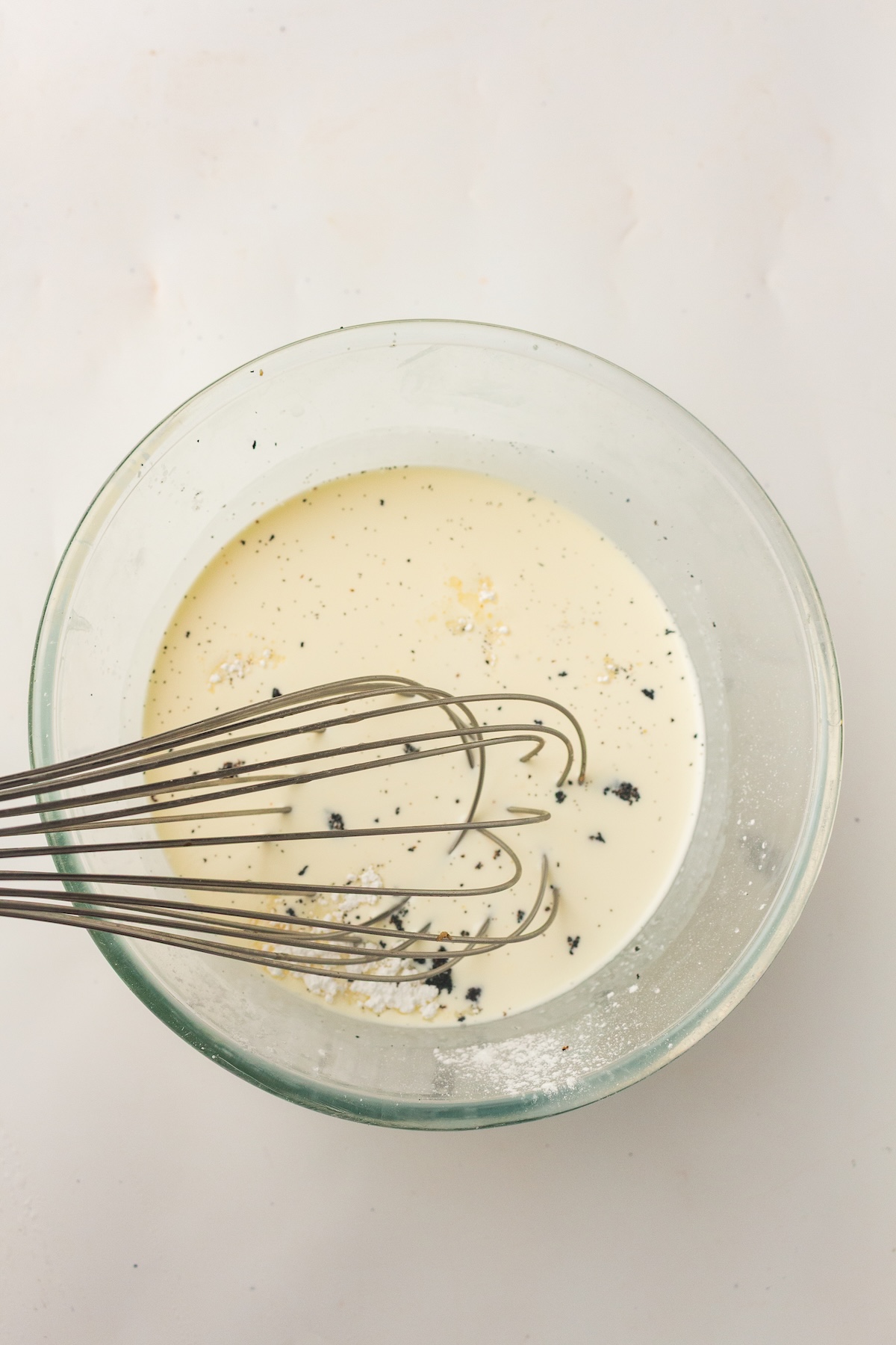 Overhead view of a clear glass bowl containing a light yellow liquid with black specks and white powder on its surface, with a metal whisk resting inside, set against a light off-white background.