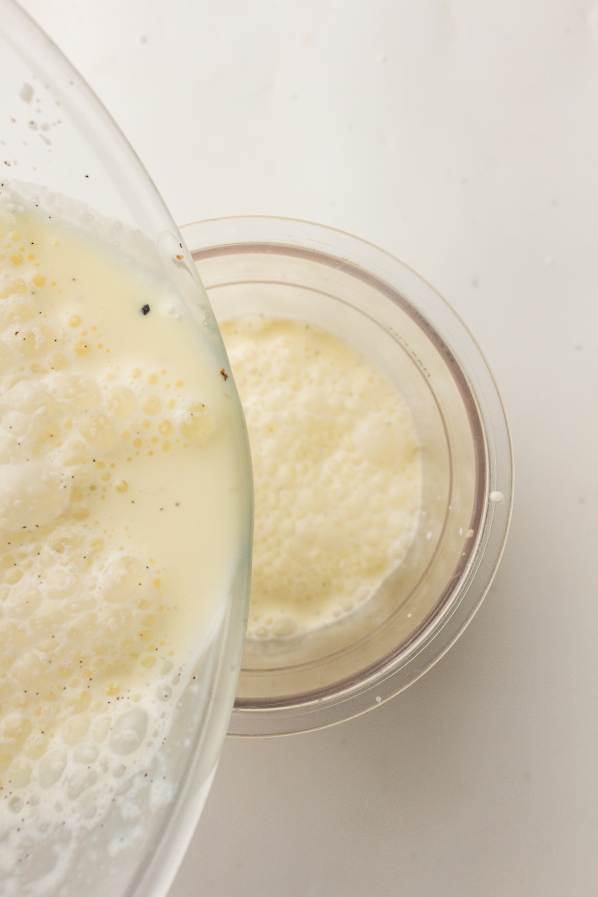 Close-up of a light yellow, frothy liquid with dark specks being poured from a large clear glass bowl into a smaller one, set against a soft off-white background.