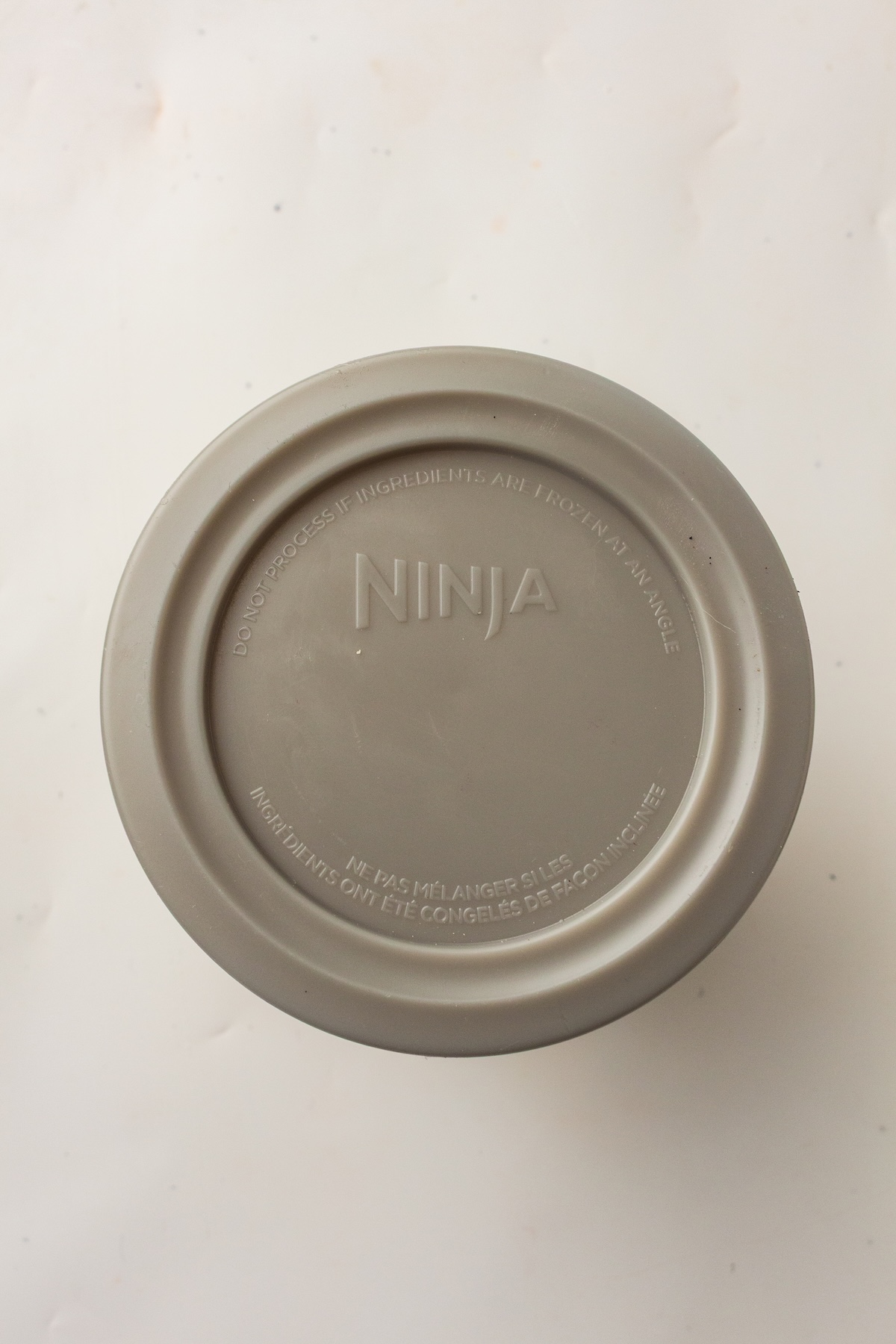 Top-down view of a circular, matte grey lid or base from a Ninja appliance, featuring embossed text with safety instructions in English and French, set on a light textured background.