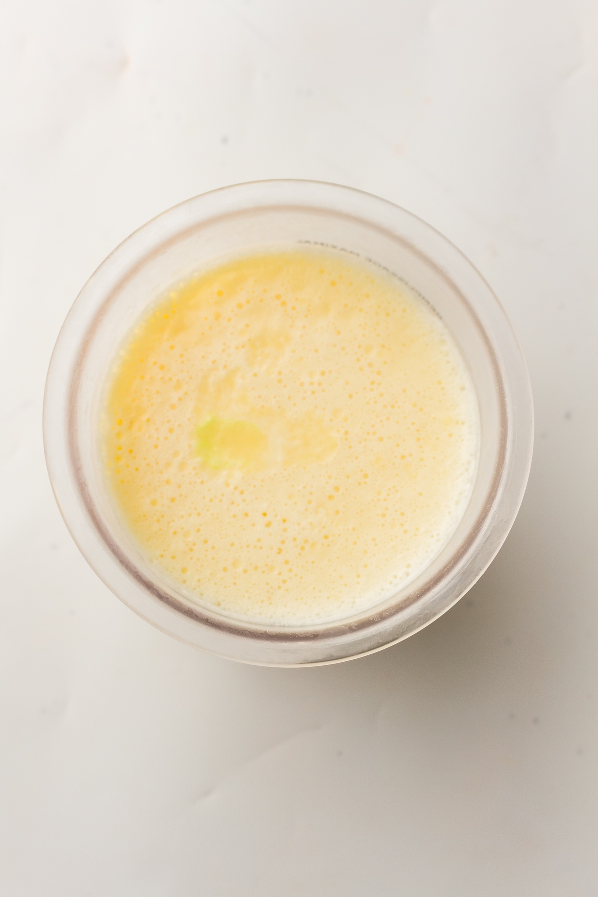 Overhead view of a translucent cup filled with a frothy, light yellow liquid, with a small brighter spot near the center, set on a plain white background.