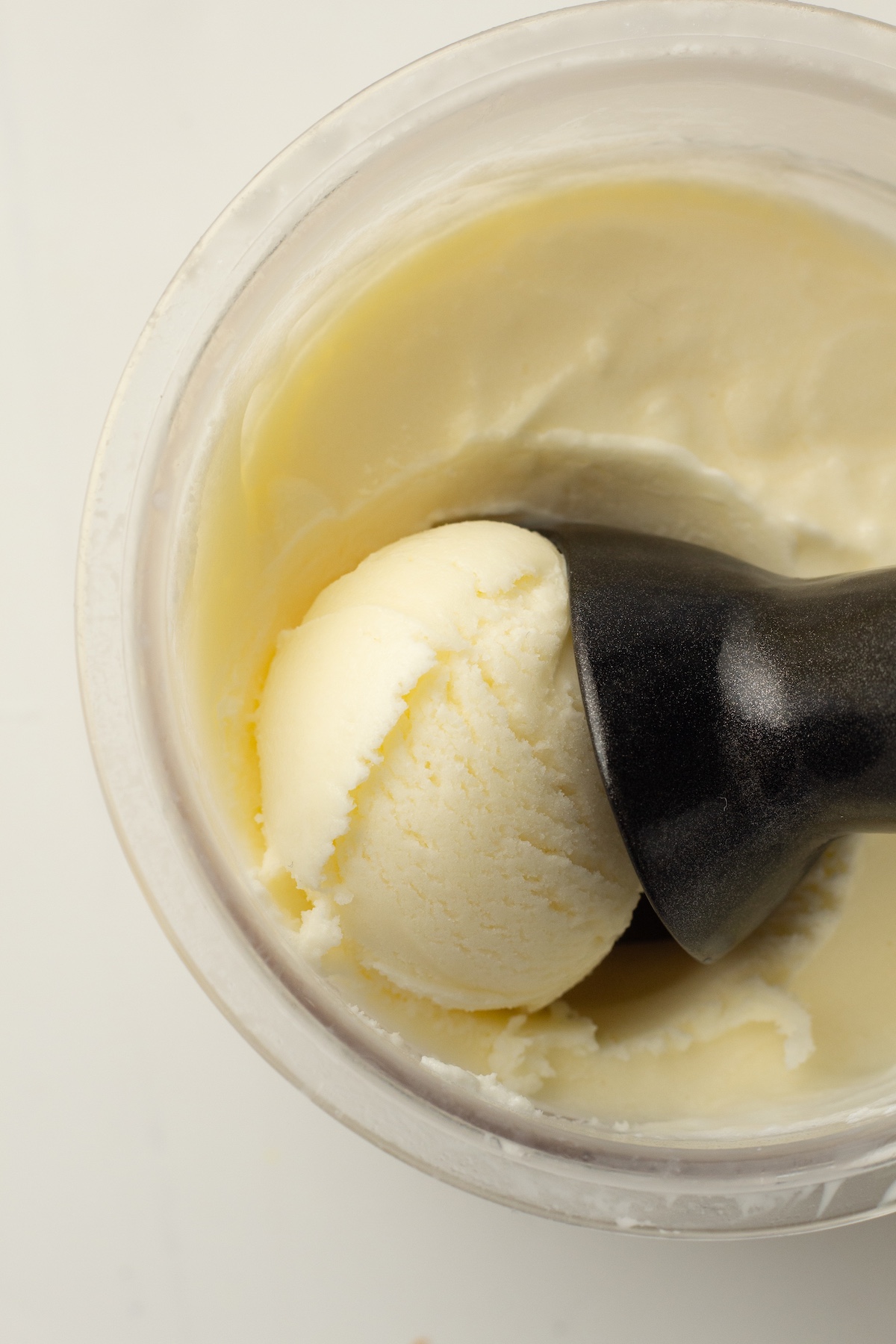 Top-down view of a clear container filled with creamy pale yellow ice cream, with a dark scoop lifting a perfectly rounded ball of ice cream.