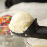 A scoop of creamy vanilla ice cream in a matte black ice cream scoop, with a blurred container of colorful sprinkles in the background.