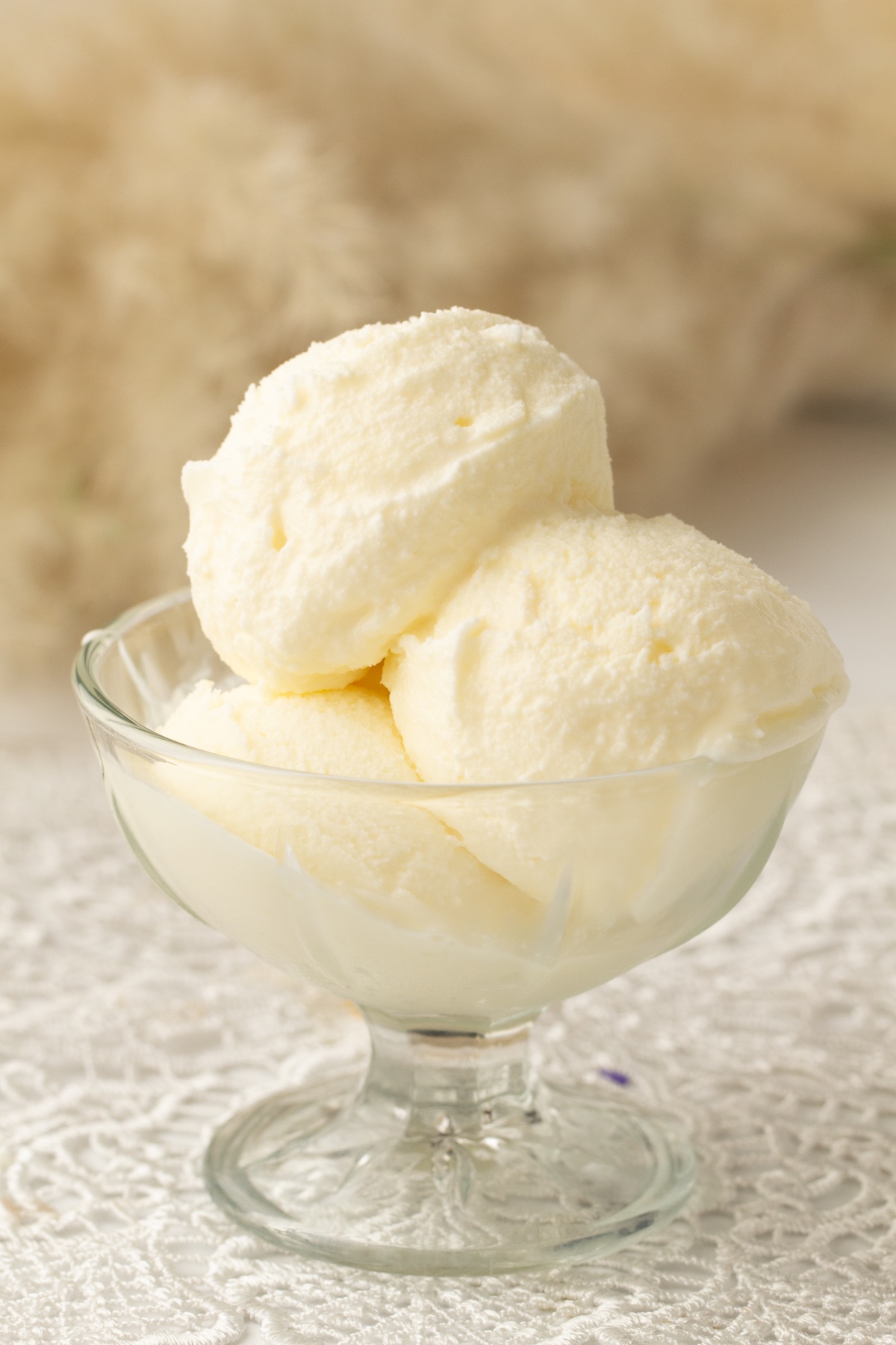 Glass bowl with scoops of creamy vanilla ice cream, placed on a white lace tablecloth, with a softly blurred warm-toned background.