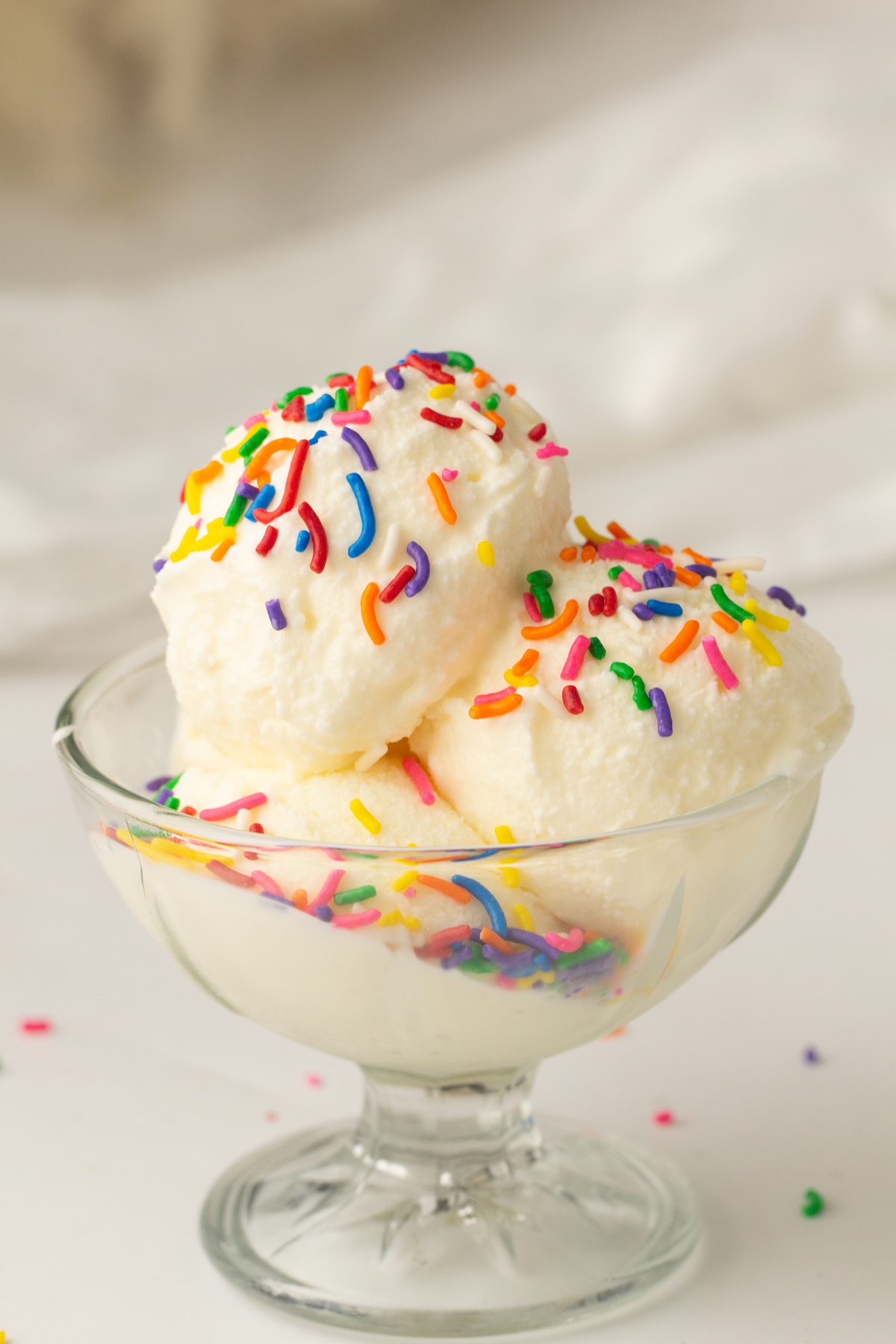 Glass sundae dish with three scoops of creamy vanilla ice cream, topped with colorful sprinkles, set against a light background.