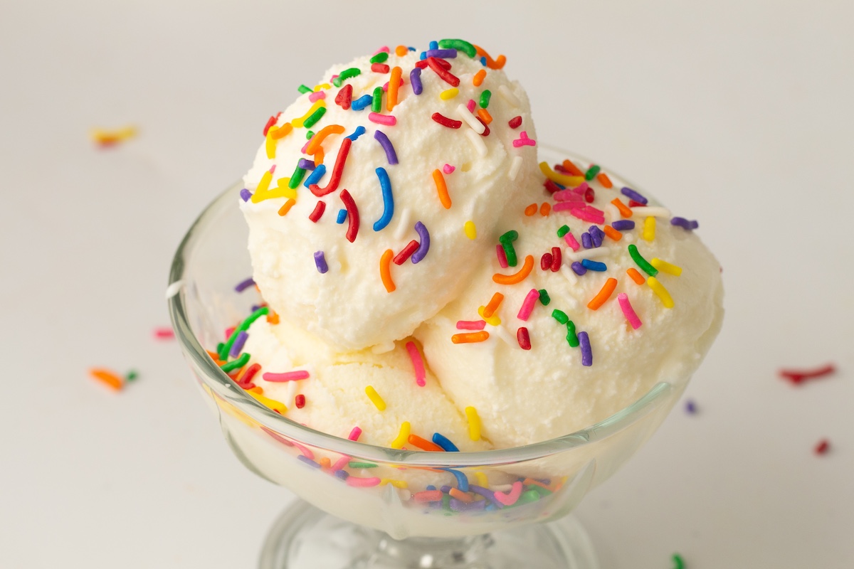 Glass sundae dish with three scoops of creamy vanilla ice cream, topped with colorful sprinkles, set against a light background.