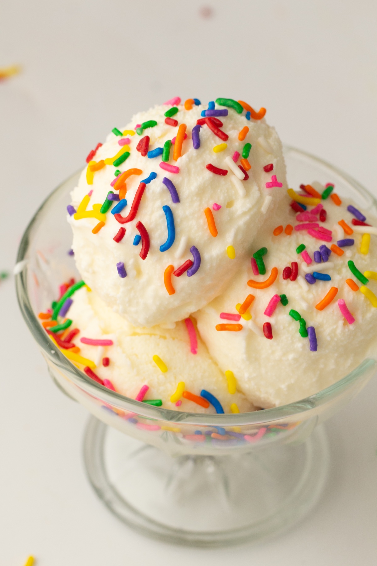 Glass sundae dish with three scoops of creamy vanilla ice cream, topped with colorful sprinkles, set against a light background.
