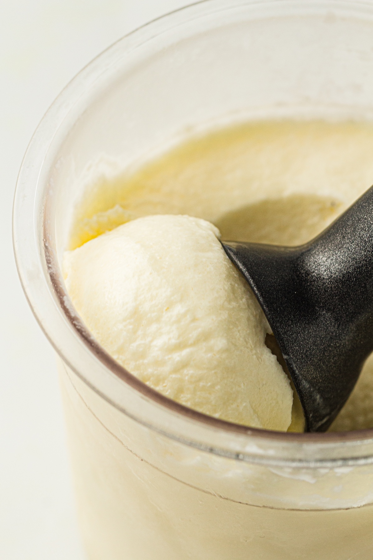 Top-down view of a clear container filled with creamy pale yellow ice cream, with a dark scoop lifting a perfectly rounded ball of ice cream.