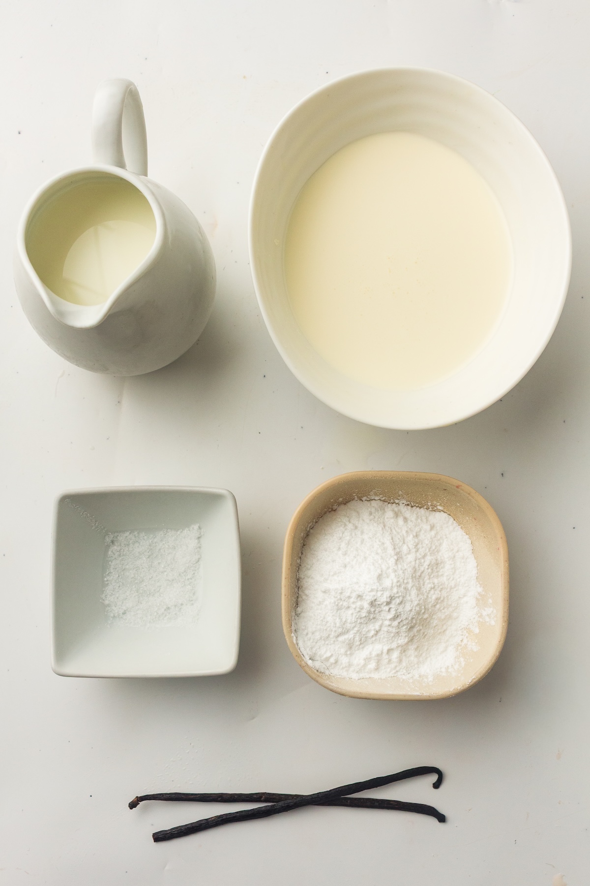 Flat lay of vanilla ice cream ingredients on a light surface, including a ceramic pitcher and bowl with cream, a small bowl of salt, a bowl of powdered sugar, and three dark vanilla beans.