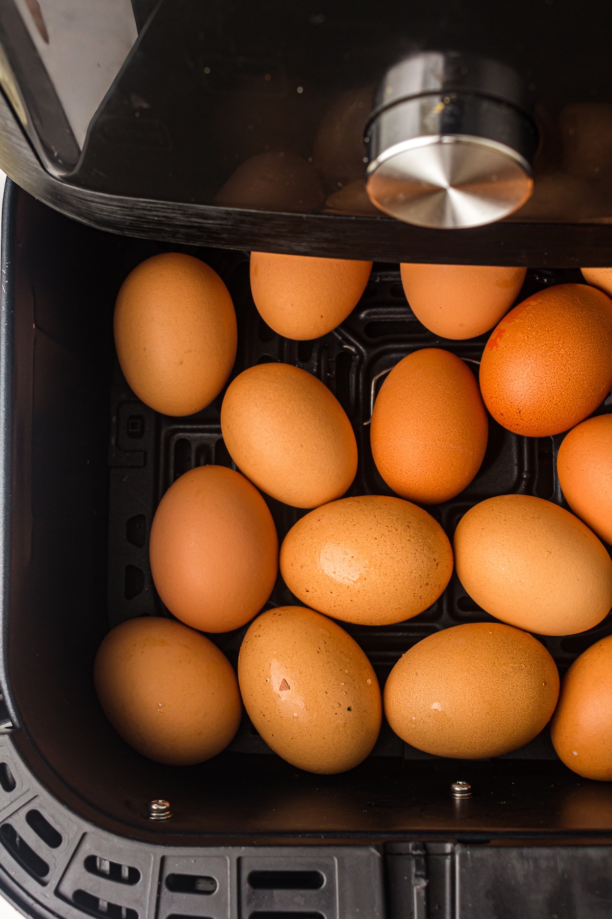 Top-down view of an air fryer basket filled with brown eggs, with the air fryer lid and a metallic control knob partially visible in the frame.