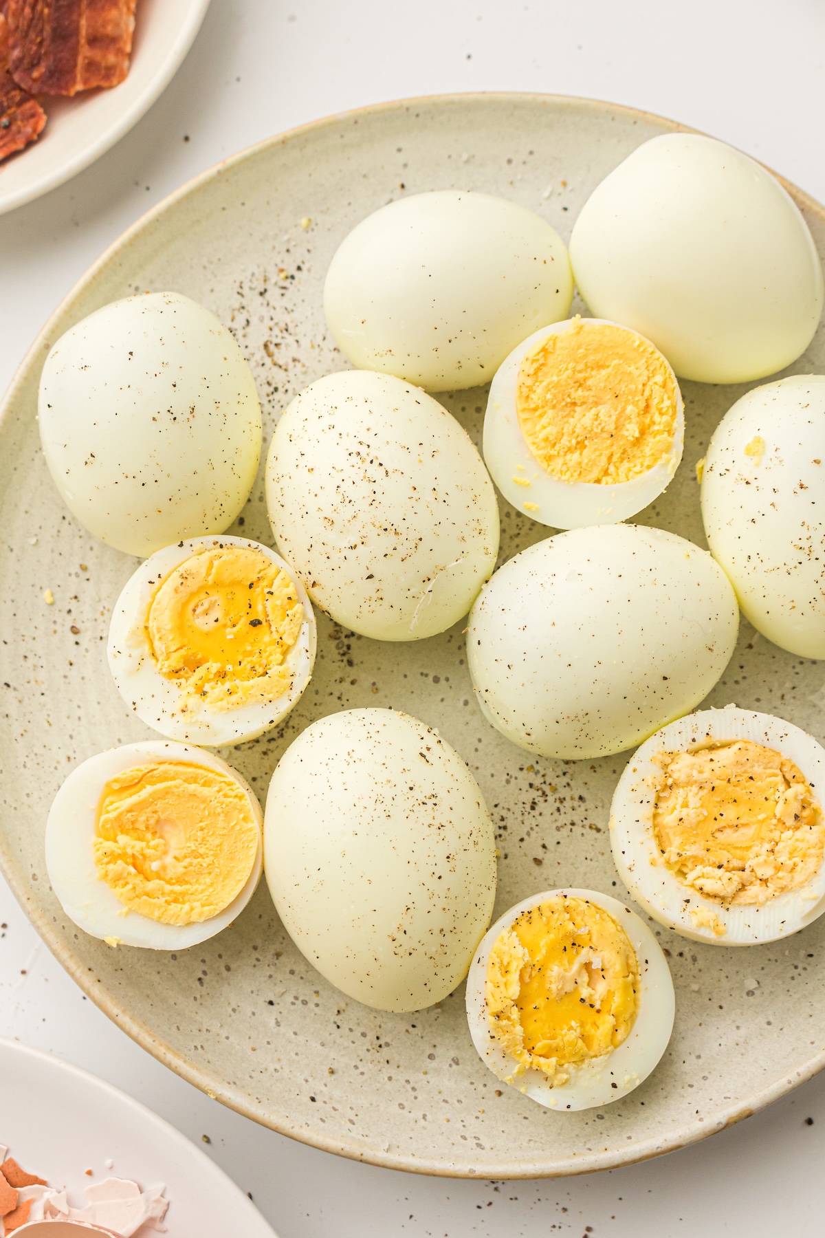 Overhead view of a plate filled with hard-boiled eggs, some whole and some halved, seasoned with black pepper, with a small dish of eggshells and a plate of bacon visible in the background.
