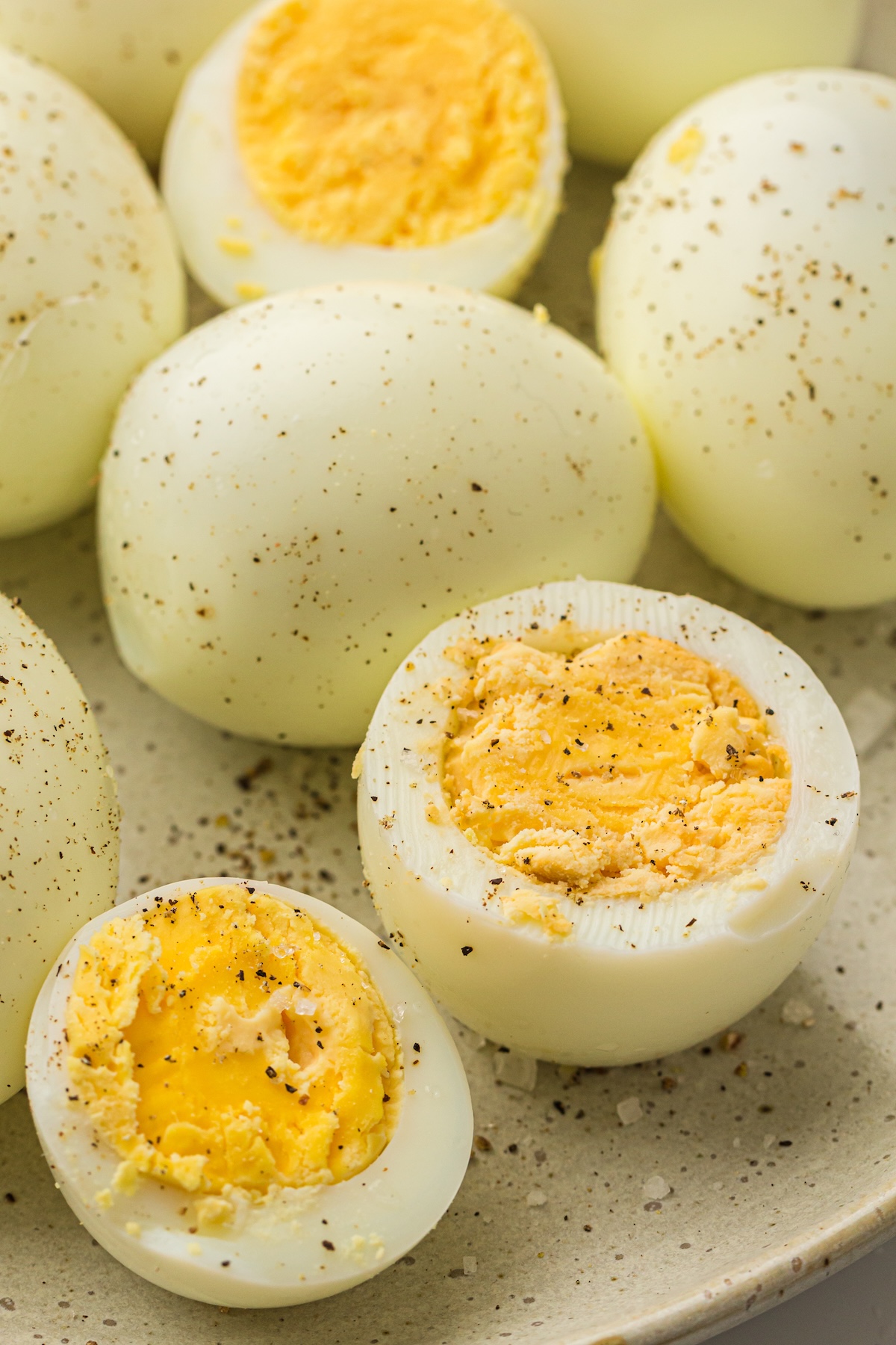 Top-down view of hard-boiled eggs on a rustic speckled plate, with a mix of whole and halved eggs seasoned with black pepper and coarse salt.
