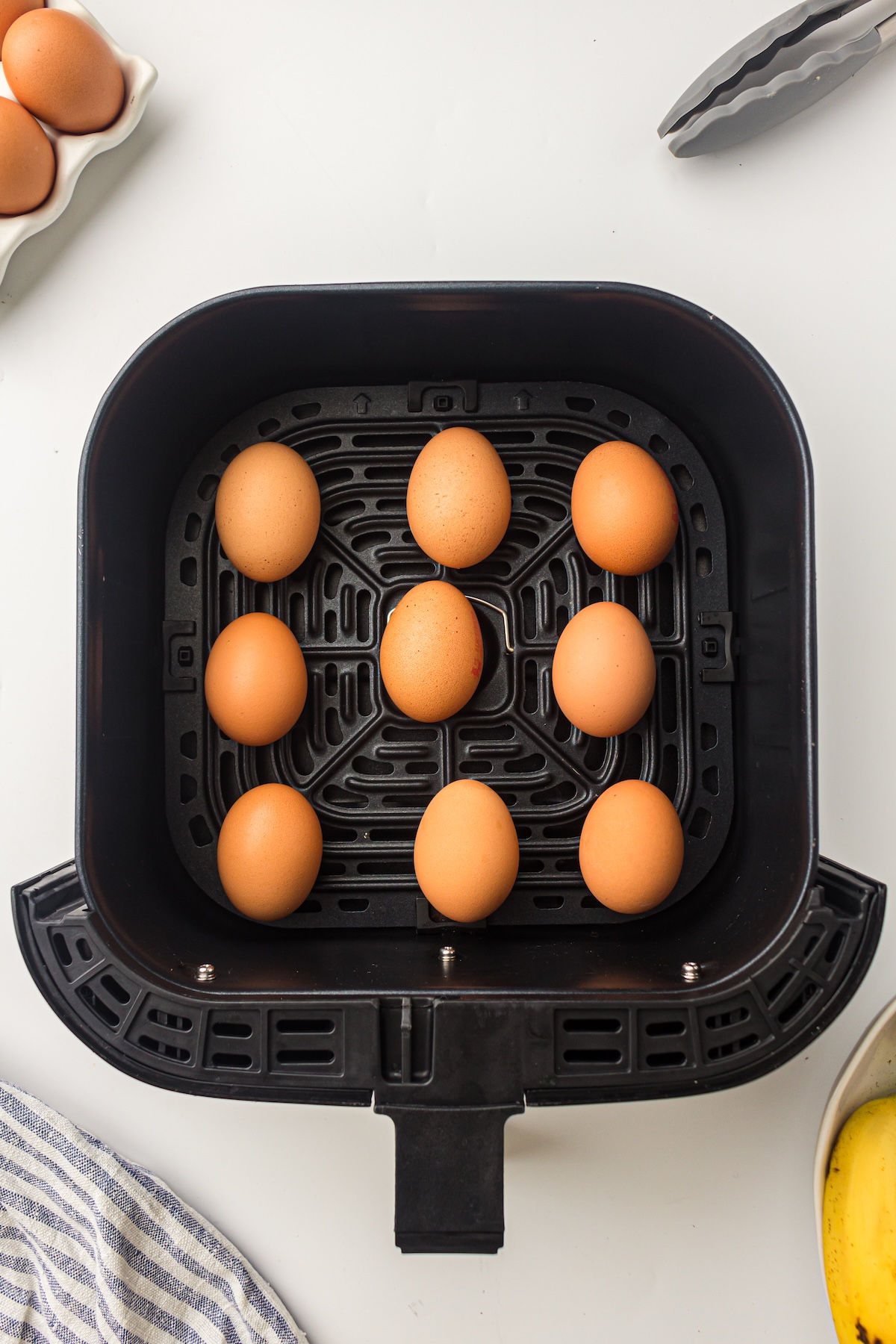 Top-down view of nine brown eggs arranged in a black air fryer basket on a white countertop, with a striped kitchen towel, tongs, and a banana in the background.
