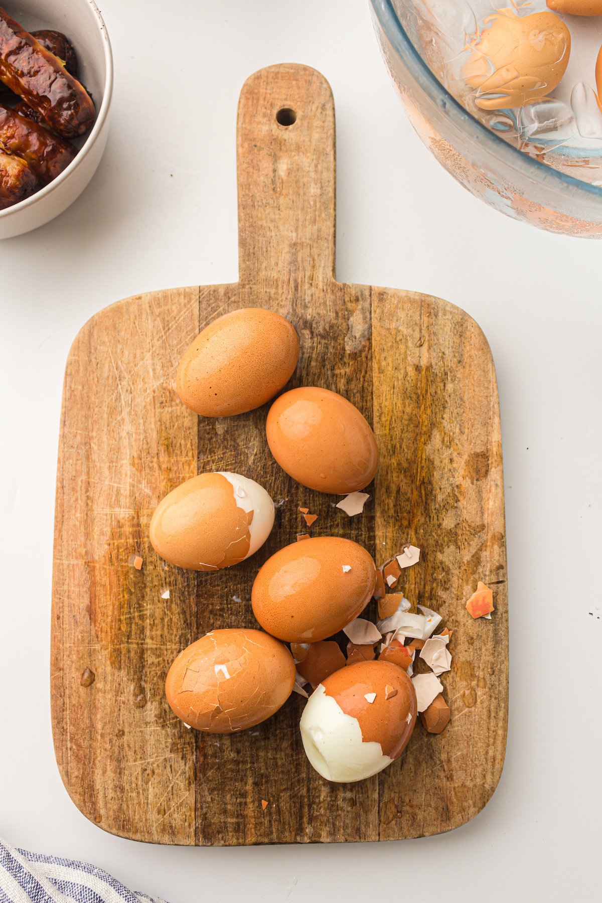 Overhead view of a wooden cutting board with five brown eggs, some partially peeled, surrounded by scattered eggshell fragments, with a bowl of sausages and a glass bowl of eggs in ice water visible in the background.