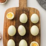 Overhead shot of a wooden cutting board with seven peeled hard-boiled eggs and two halved eggs showing bright yellow yolks, surrounded by a carton of unpeeled eggs, a striped cloth, and scattered eggshells.