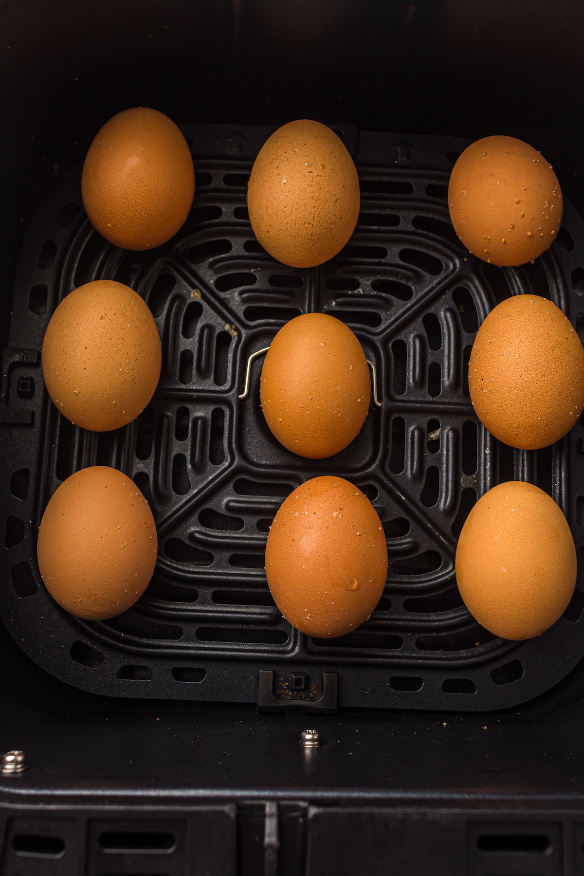 Overhead shot of nine brown eggs arranged in a black air fryer basket with a perforated bottom, ready for cooking.