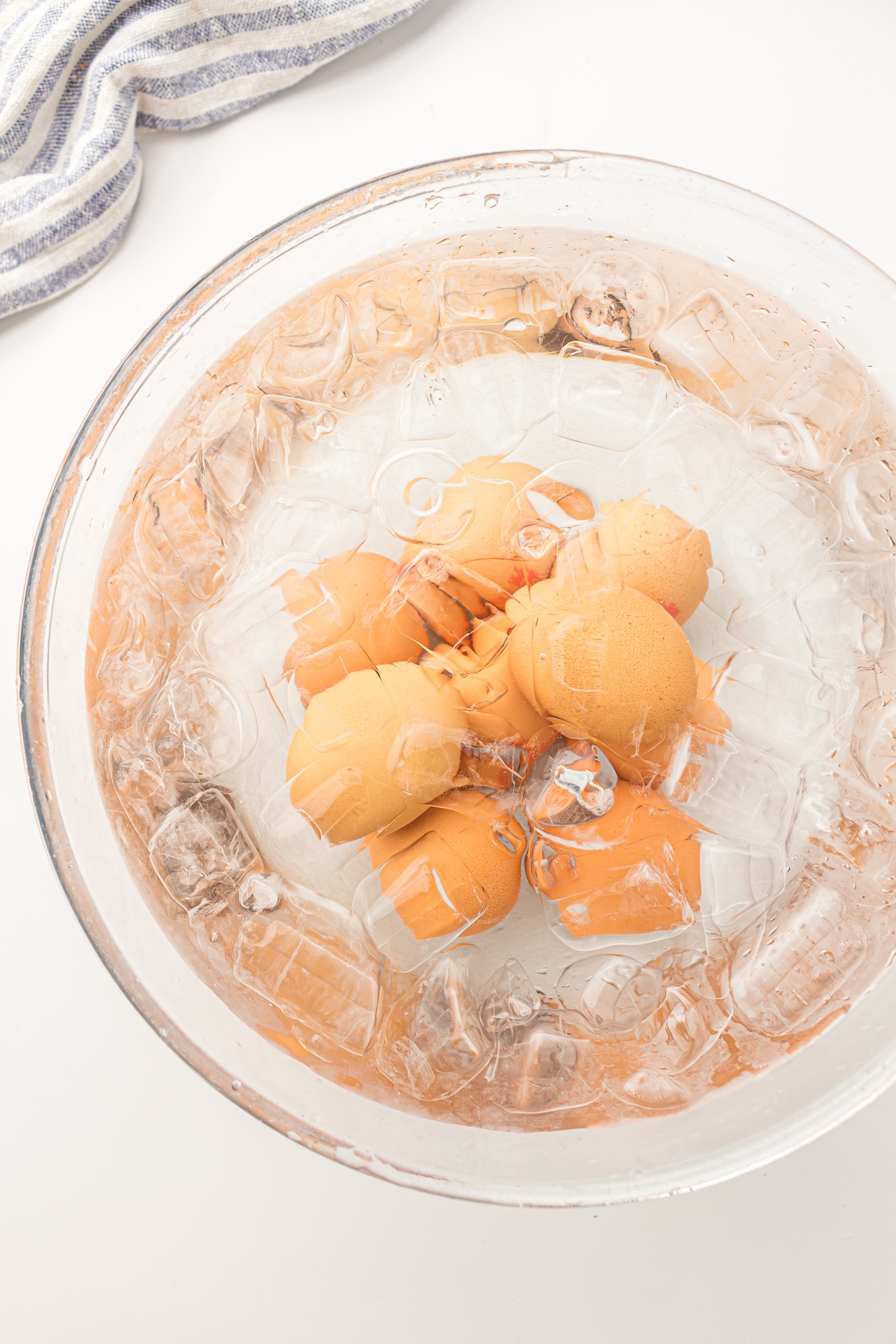 Top-down view of a clear glass bowl filled with ice water and several light brown eggs, surrounded by floating ice cubes, resting on a white surface with a striped kitchen towel in the corner.