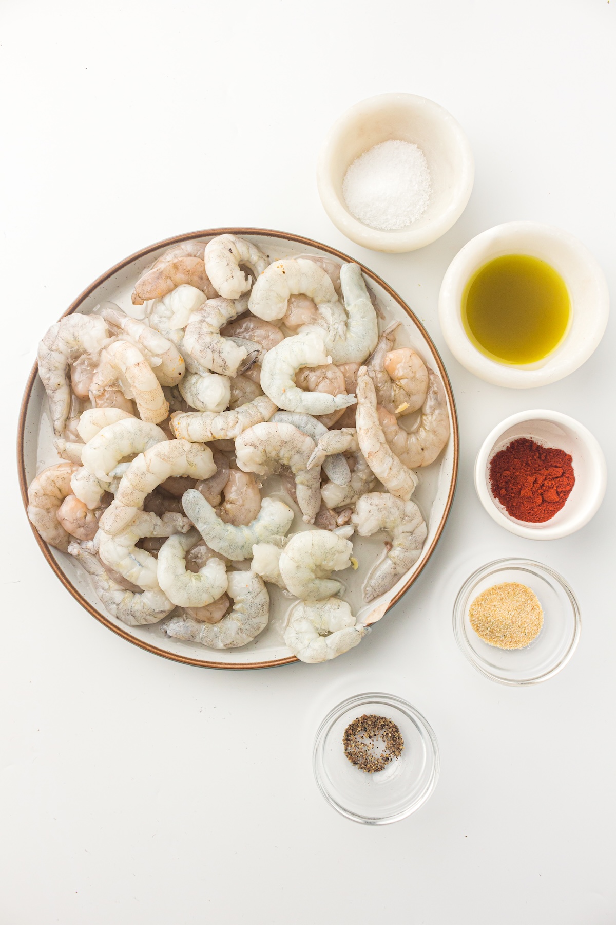 Flat lay of raw shrimp on a ceramic plate surrounded by small bowls of salt, olive oil, paprika, garlic powder, and black pepper, set against a clean white background.