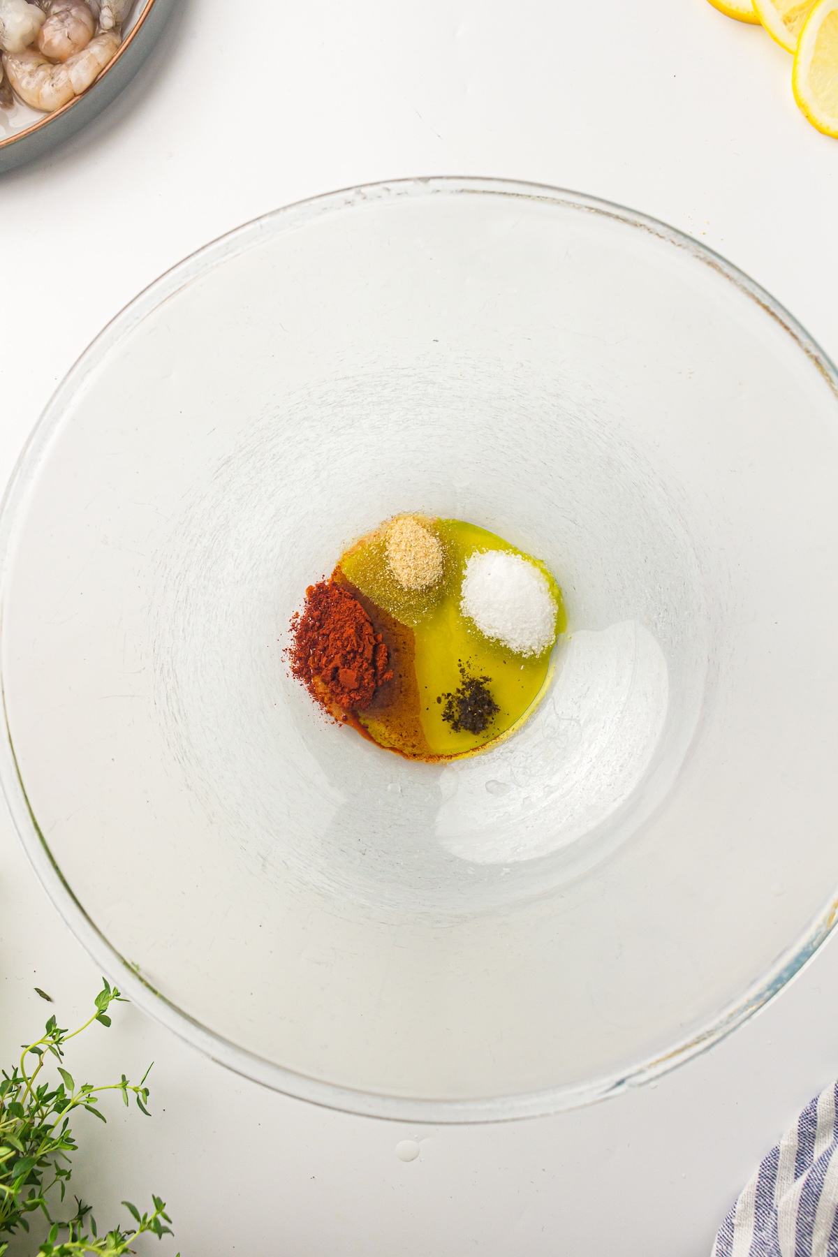 Top-down view of a glass bowl with olive oil and spices, including paprika, garlic powder, salt, and black pepper, ready to be mixed for a shrimp marinade.