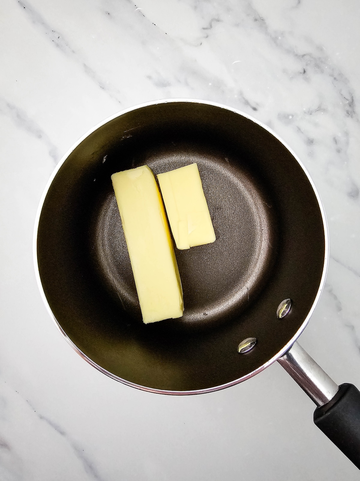 Overhead view of a dark non-stick saucepan containing two solid sticks of butter, placed on a white marble countertop.