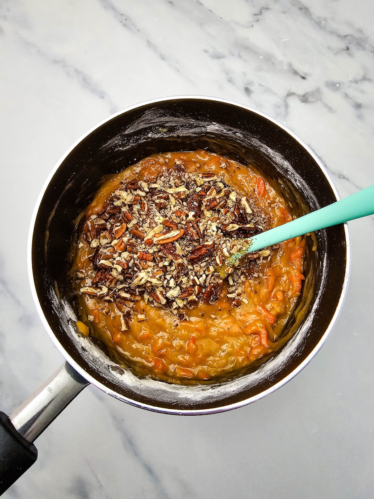 Top-down view of a dark saucepan containing a thick orange-brown batter topped with chopped pecans and darker sprinkles, with a light turquoise silicone spatula resting in the mixture, on a white marble countertop.