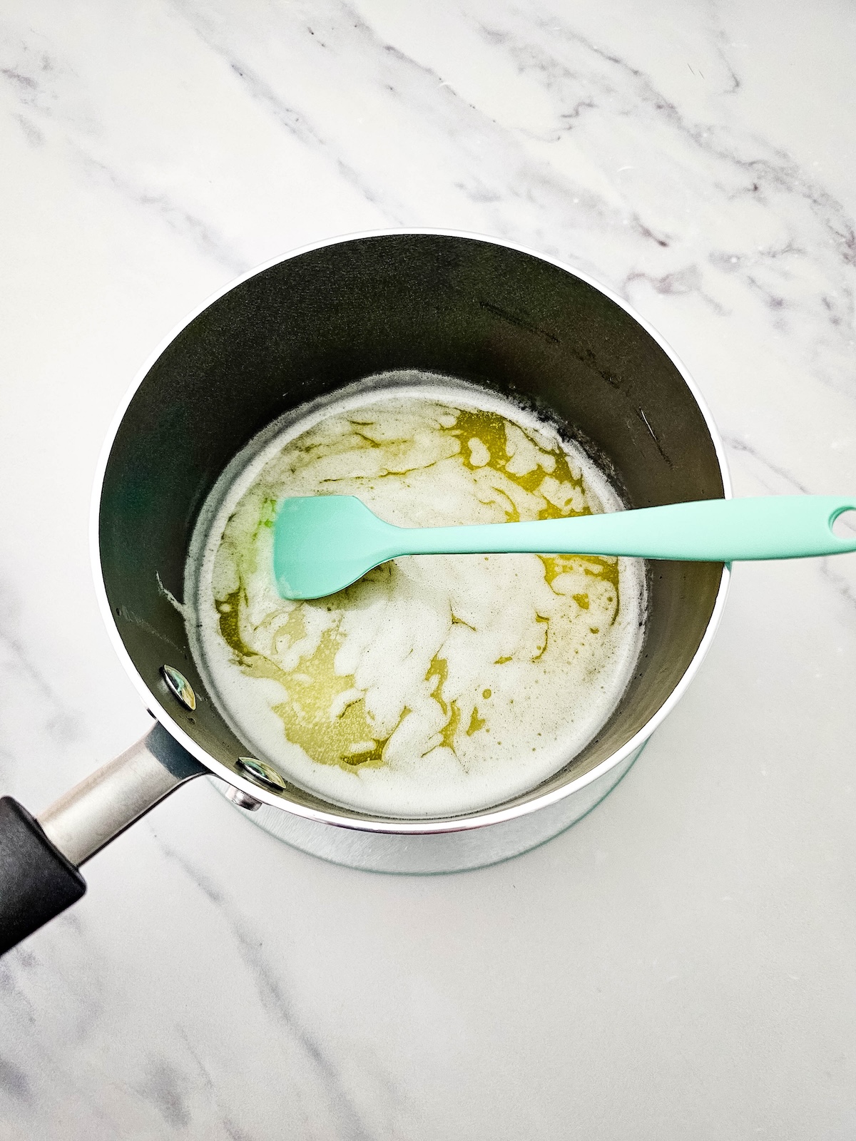Overhead view of a dark saucepan with melted butter bubbling and frothing, accompanied by a teal silicone spatula resting inside, on a white marble surface.