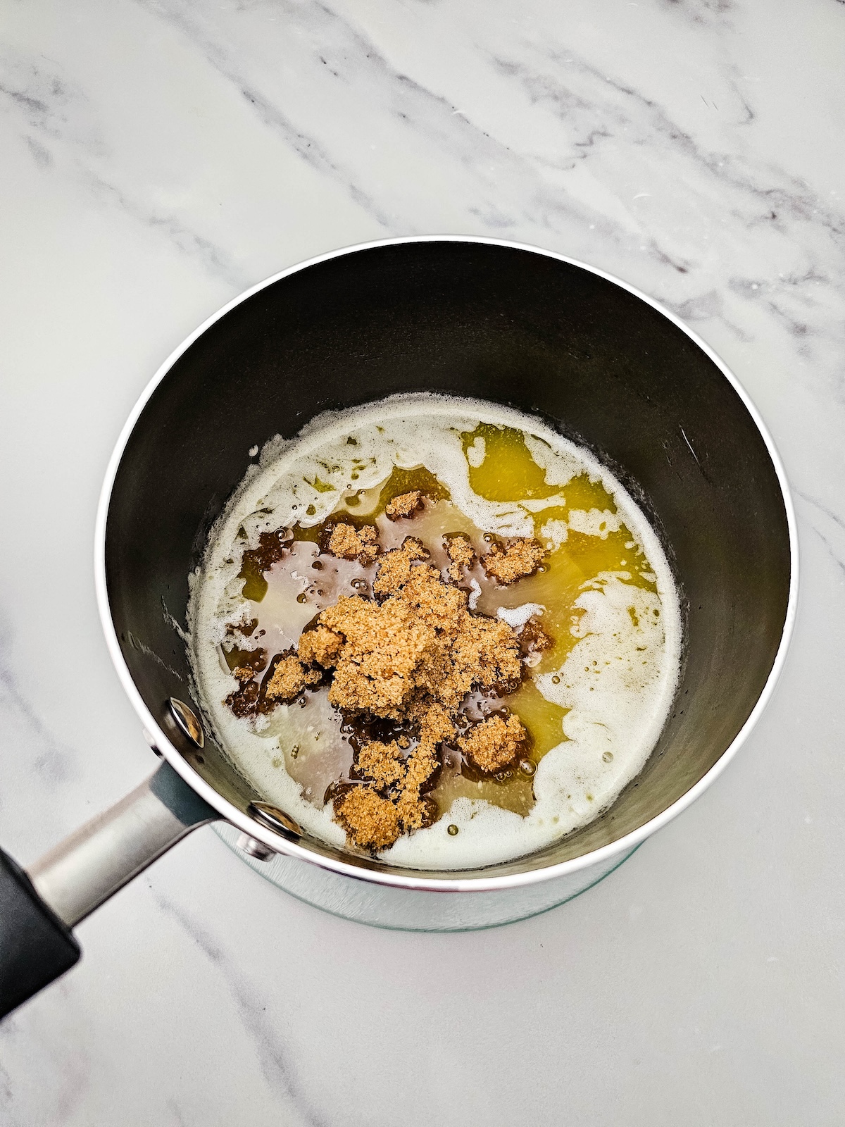 Overhead view of a dark saucepan containing melted butter mixed with clumps of brown sugar, creating a golden, glossy mixture, on a white marble countertop.
