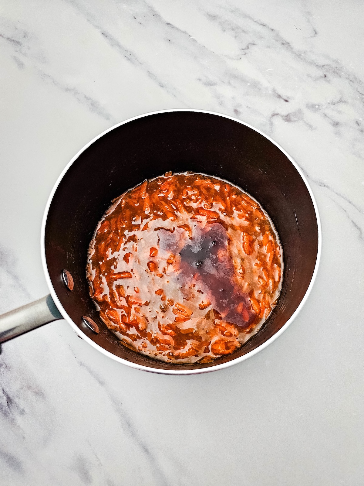Overhead view of a black saucepan with a thick reddish-orange mixture, featuring visible shredded carrots and a darker unincorporated liquid spot, on a white marble countertop.