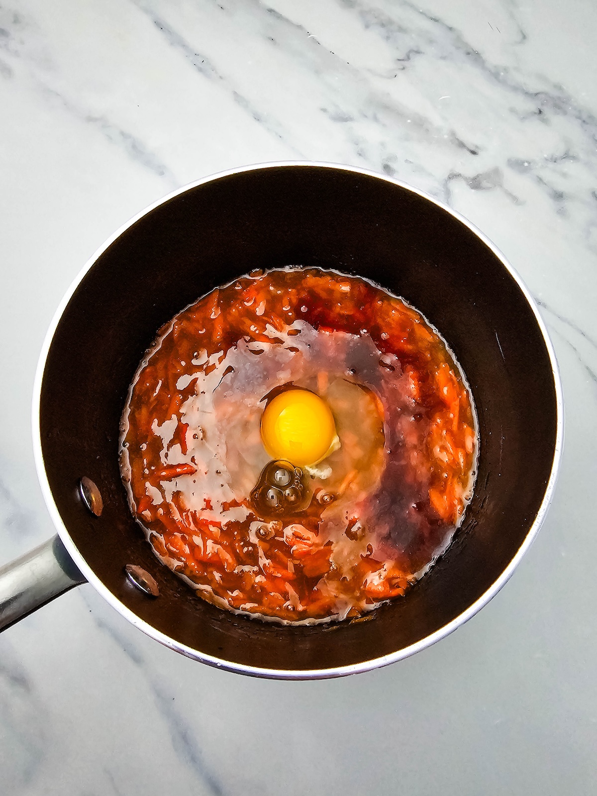 Overhead view of a dark cooking pot containing a reddish-brown mixture with a raw egg cracked in the center. The yolk is vibrant orange, surrounded by clear egg white, on a white marble countertop.