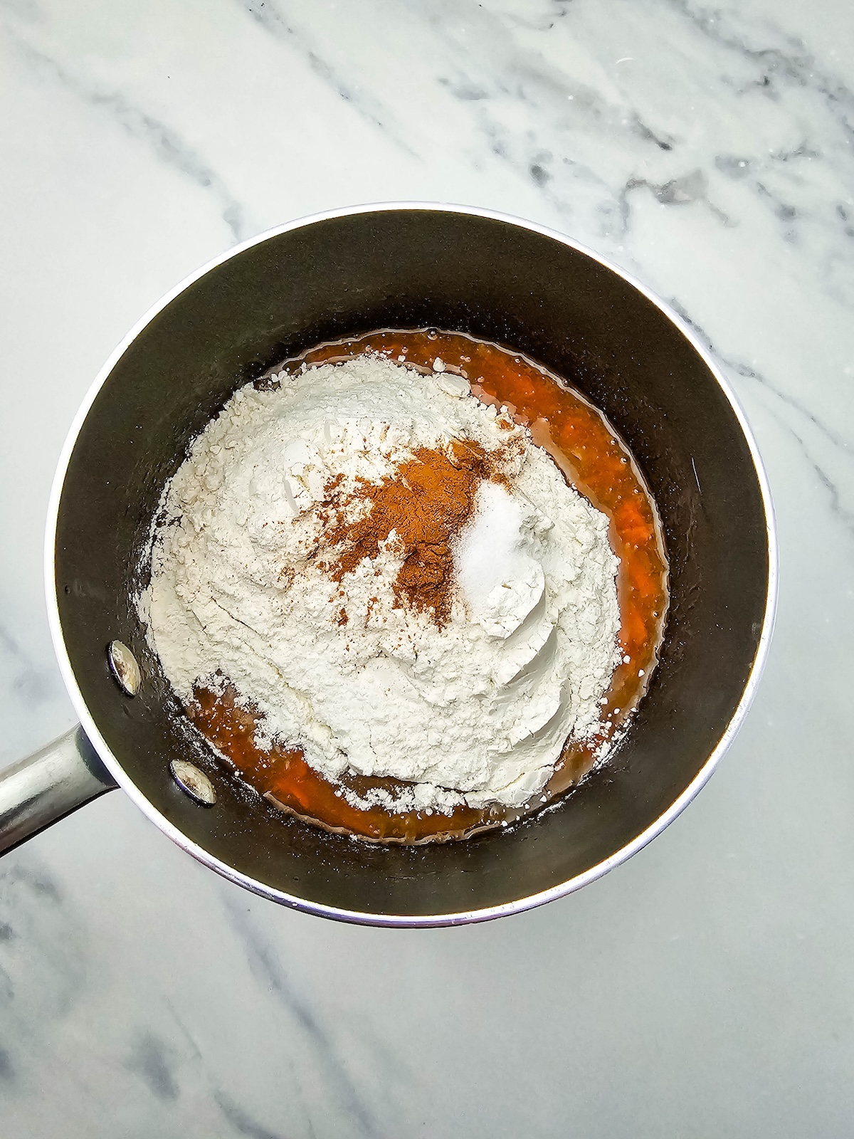 Overhead shot of a dark saucepan containing an amber liquid base topped with a mound of white flour, a sprinkle of reddish-brown cinnamon, and a smaller pile of white powder, likely salt or baking soda, on a marble countertop.